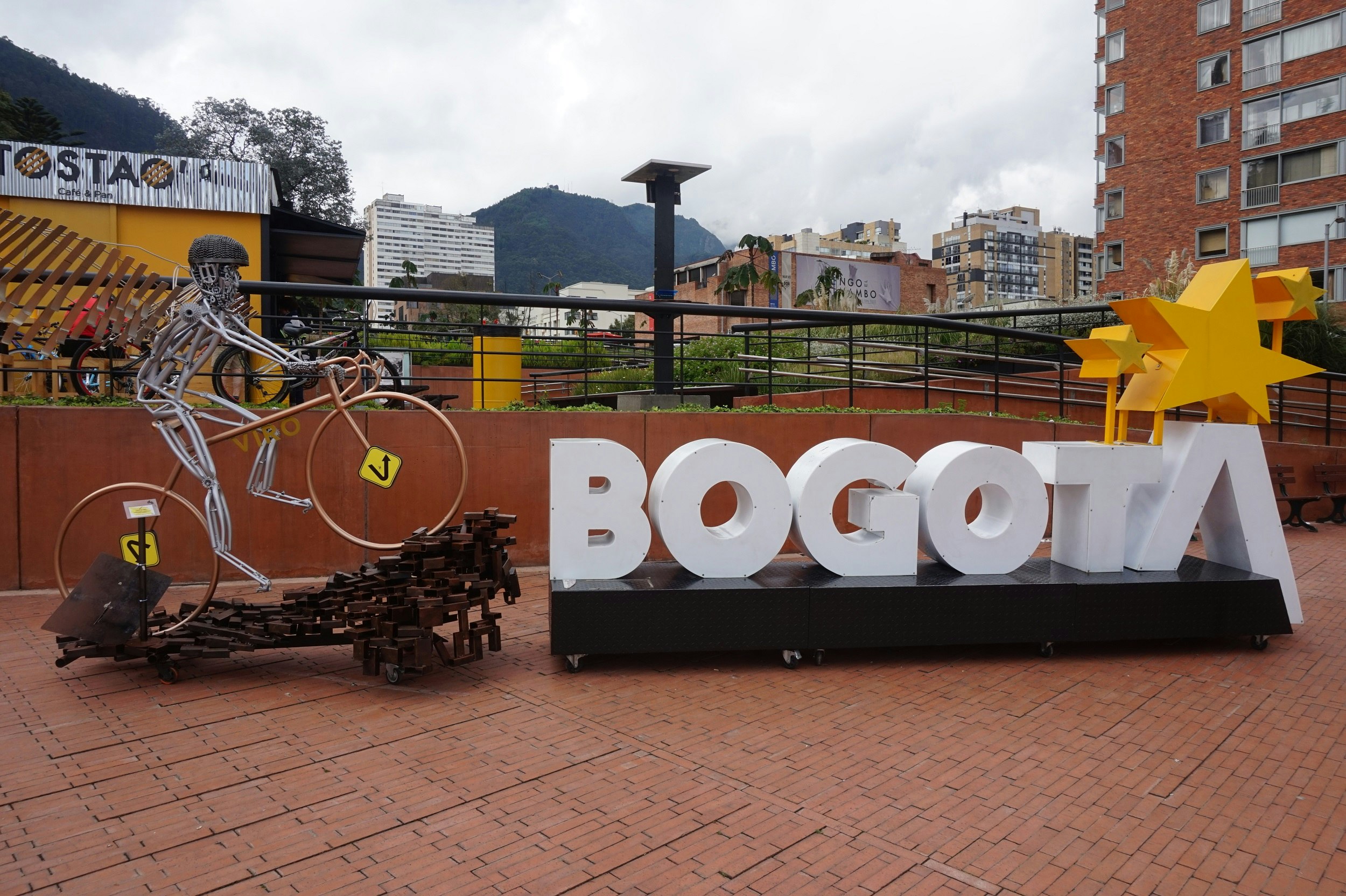 A moveable (on coasters) display for Cyclovia; to the left is a life-size statue of a cyclist climbing a hill (made of scrap metal), while to the right is a long black box with the words Bogota spelled out with large white block letters (three gold stars rise above the T and A at the end.)