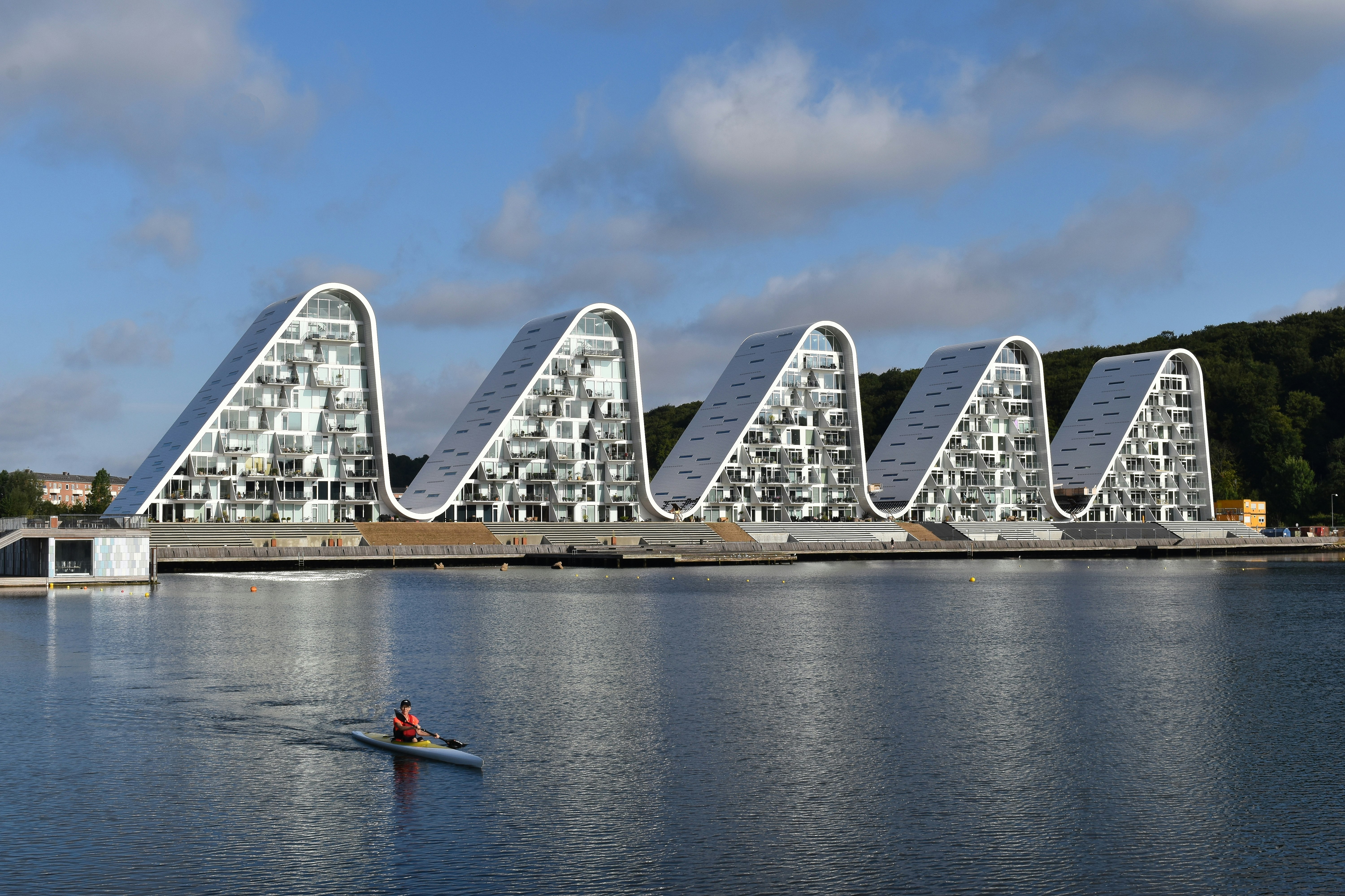 Five white, wave-shaped buildings stand at the water's edge in Vejle, Denmark; there's a lone kayaker paddling in the harbour.