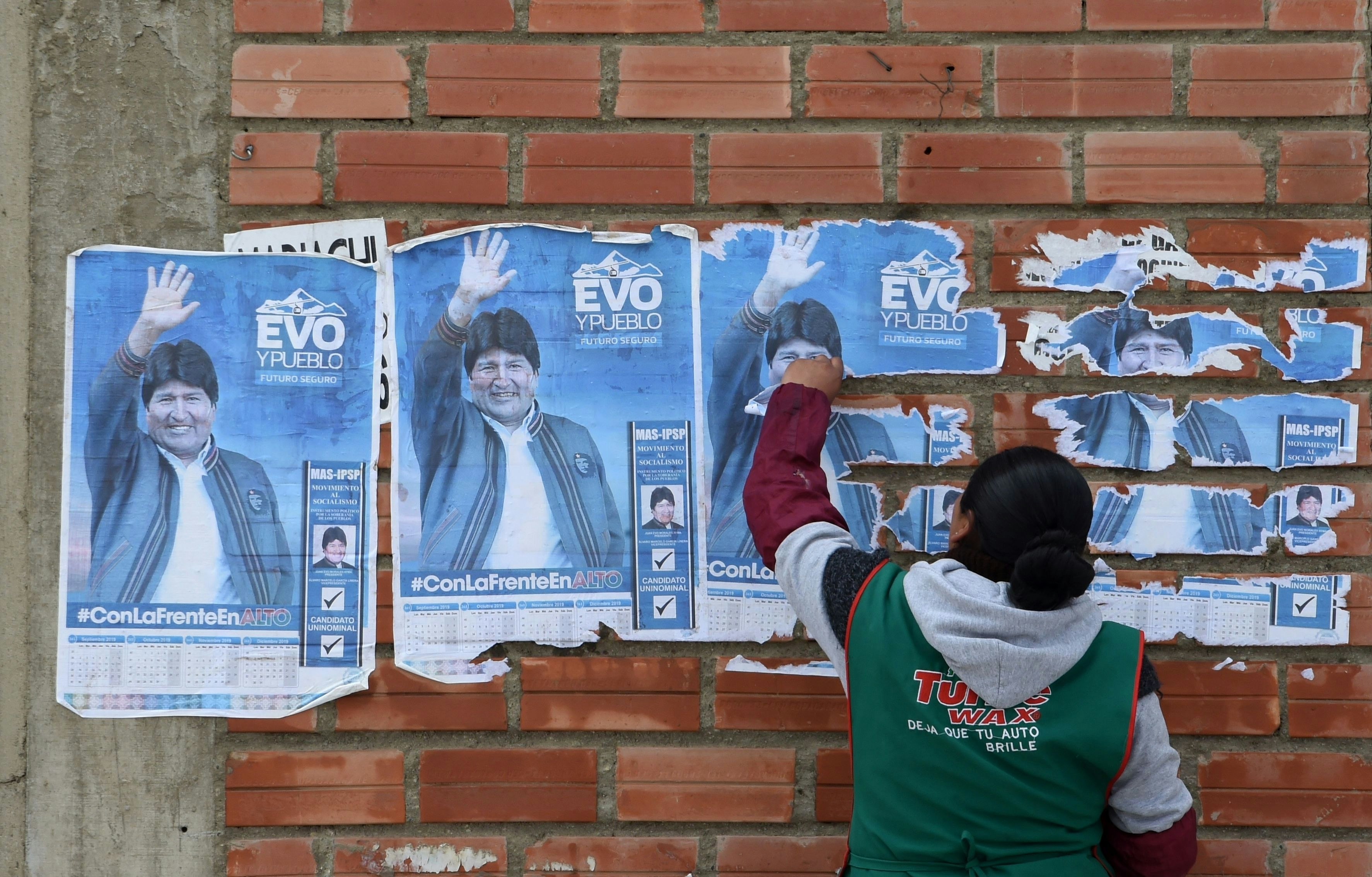 A woman peels posters of former Bolivian president Evo Morales off of a red brick wall. 