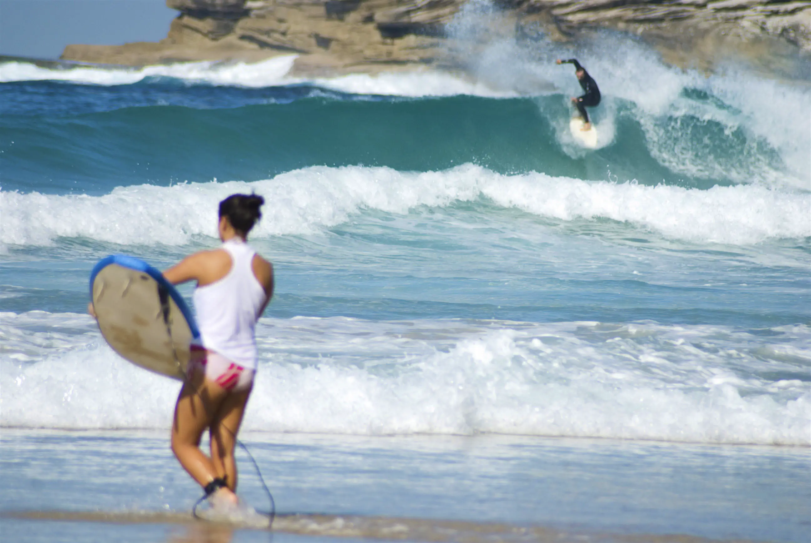 bondi-beach-LPI-25412-79.jpg A woman in a swimsuit walks into the surf holding a board, while a wetsuit-clad surfer rides a large wave in the distance.