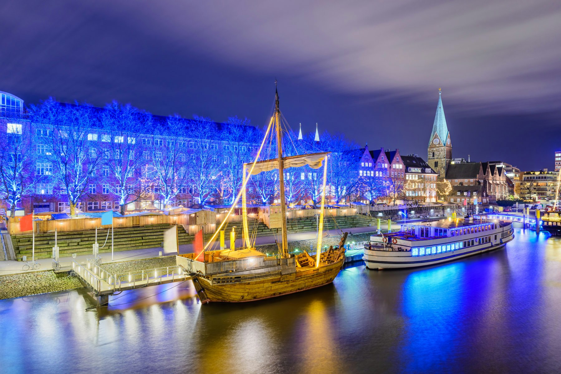 Ships on the river in Bremen lit up. In the background are a row of houses completely illuminated by blue lights.