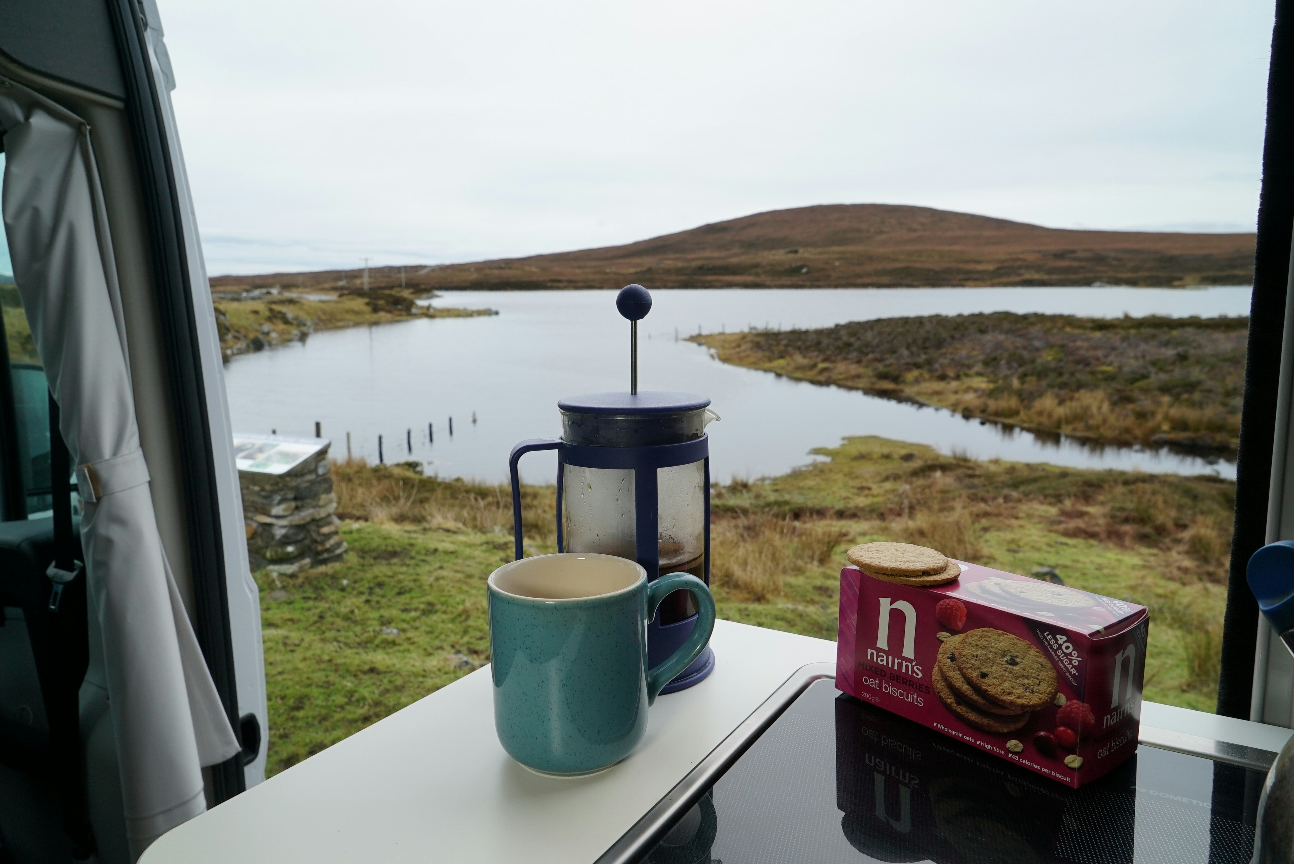 A mug of coffee and accompanying biscuits stand on a table of a camper van with the door open behind it, revealing a scenic view of a lake and green mountains.