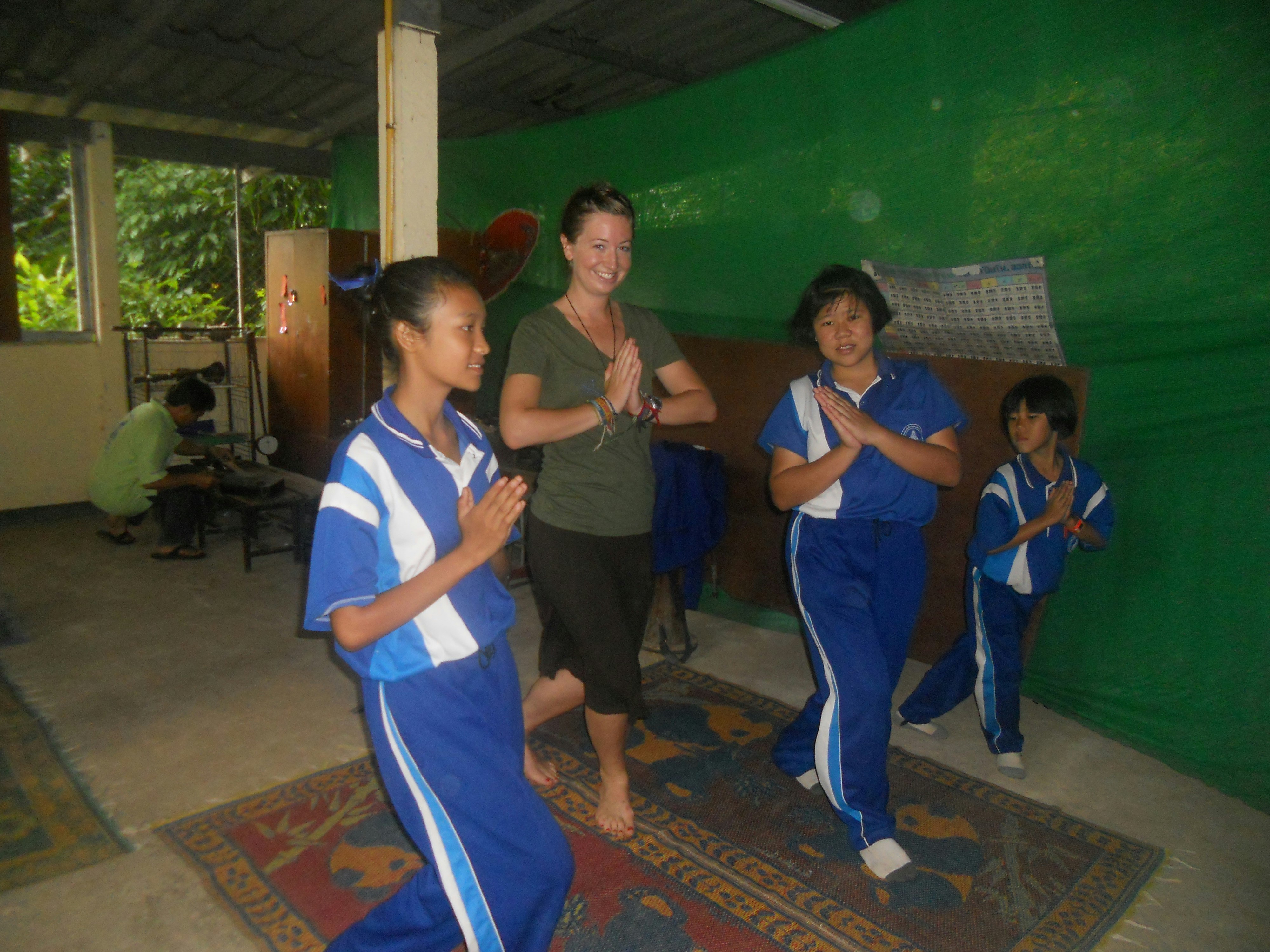 Environmental writer Britany Robinson stands with three Thai girls of varying ages. The girls are in blue track suits with their palms pressed together. Britany is in a green top and black pants striking a similar pose.