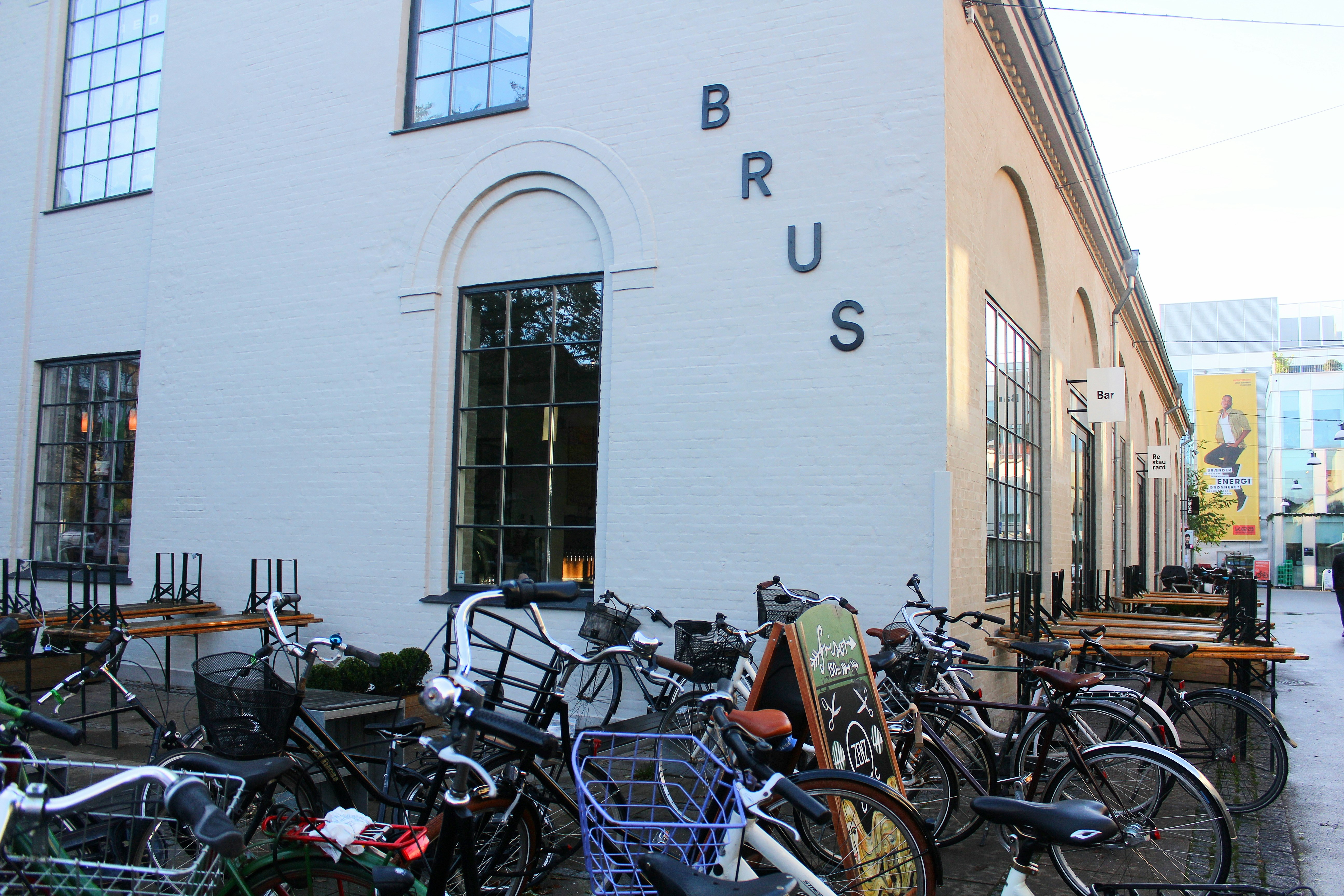 The exterior of Brus microbrewery in Copenhagen; many bikes are parked outside the simple whitewashed building that has 'BRUS' spelled out in individual letters on the side.