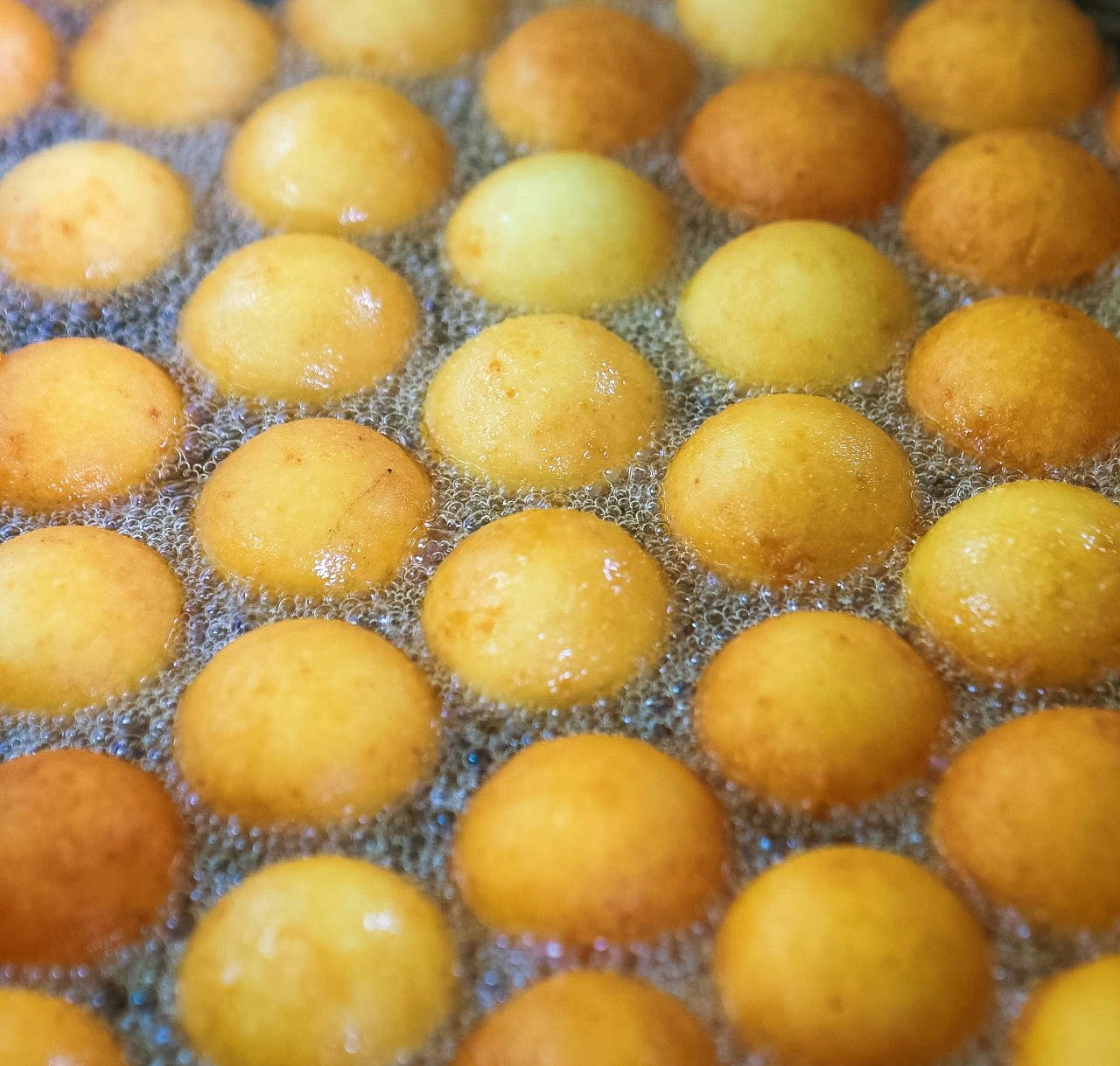 Piping hot bunuelos sit in a bath of bubbling oil. Each bunuelo is a round, pale yellow sphere (some darkening into brown) surrounded by tiny white and clear bubbles.