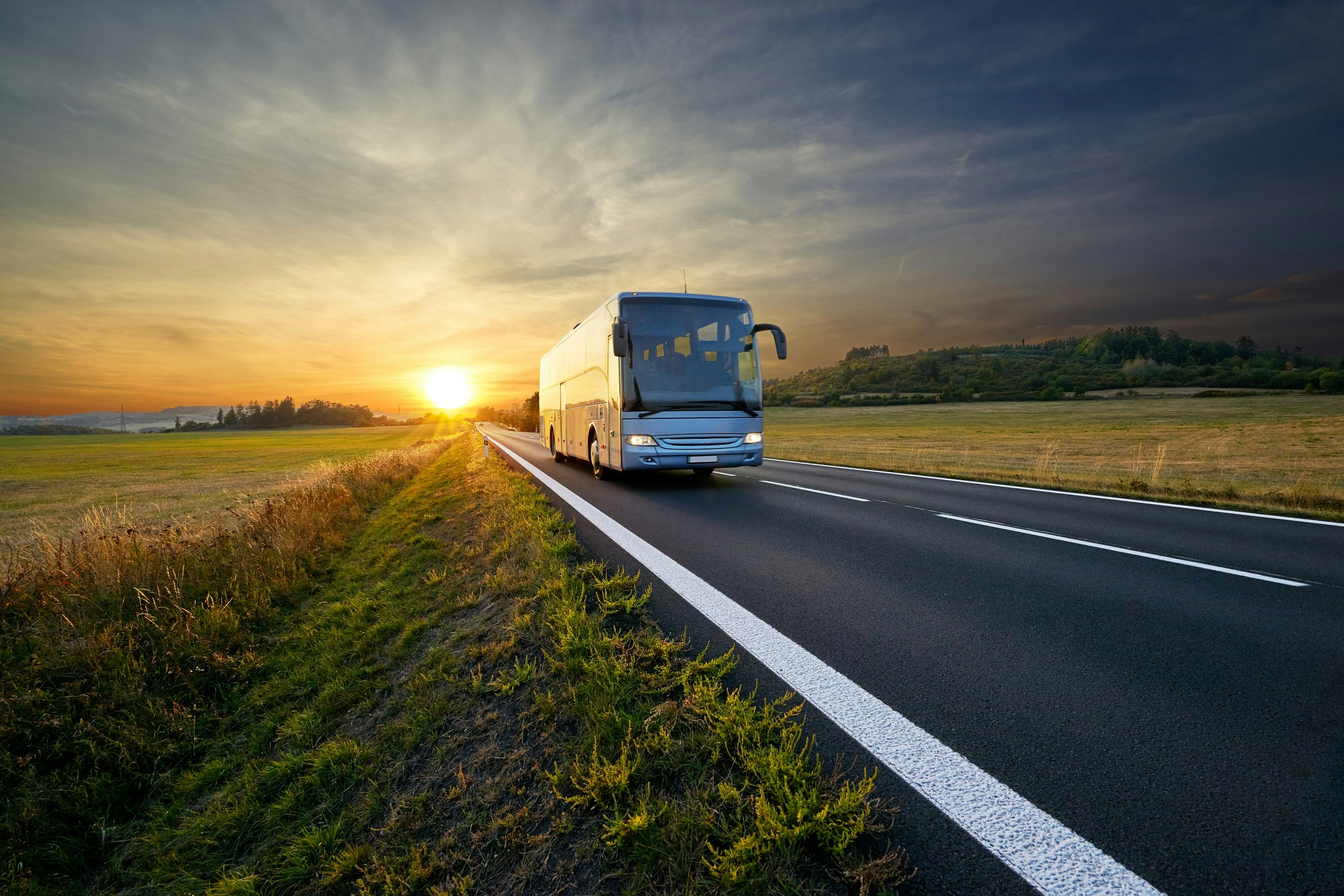 A large coach driving along a road in a rural region with the sun setting in the background