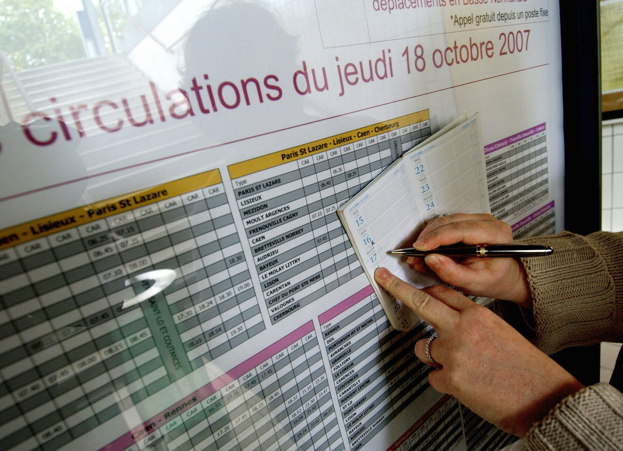 A pair of hands with a wedding band and a tan cable knit sweater visible, uses a black and gold fountain ballpoint pen to copy times off a large bus schedule at a station or stop into a small personal diary.