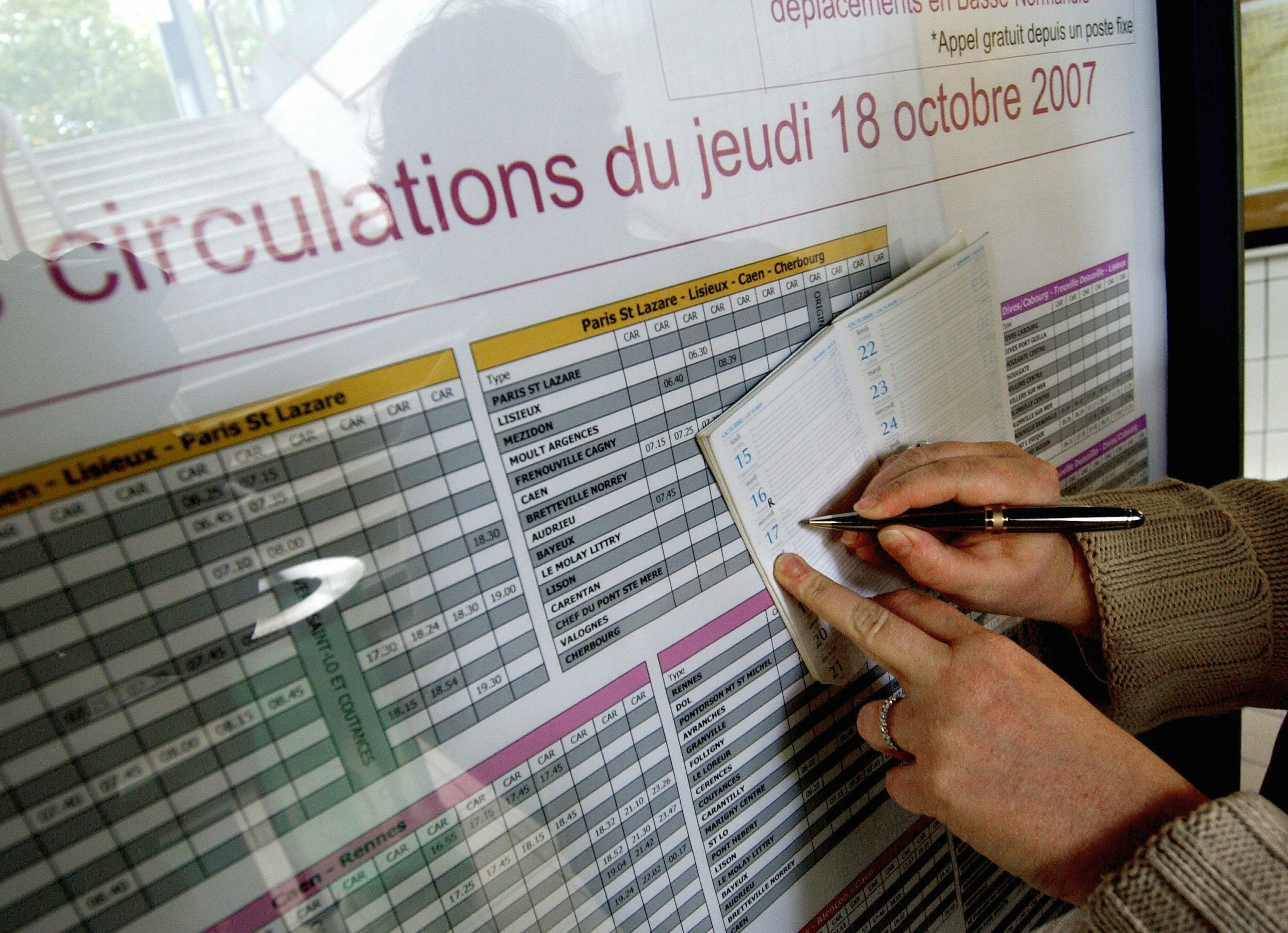 A pair of hands with a wedding band and a tan cable knit sweater visible, uses a black and gold fountain ballpoint pen to copy times off a large bus schedule at a station or stop into a small personal diary. 