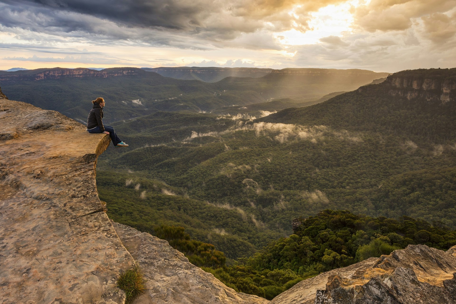 Woman sitting on a rocky ledge overlooking the Blue Mountains National Park during sunset; in the bottom of the valley are fluffy clouds floating over the trees.