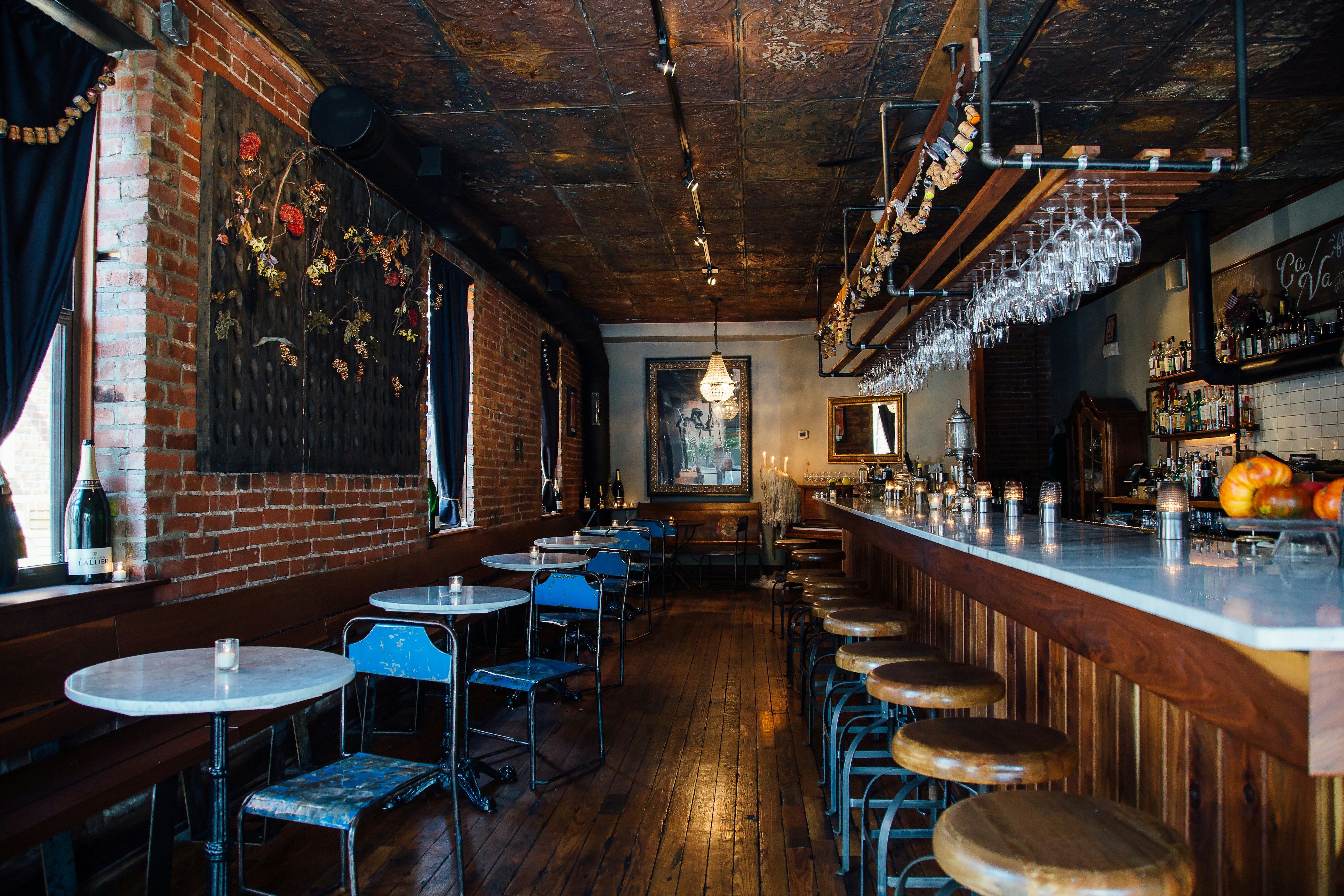 The warm, dark interior of ça va champagne bar in Kansas City is made up of tin ceiling panels with a rich bronze patina, worn wide-plank floor boards with a deep finish, a wooden bar with a marble top, garlands of corks in champagne caages, wood and metal industrial stools, and marble and iron cafe tables. Dark floral art hangs on the walls and simple crystal chandeliers and candle votives glow. In the far background, a pedestal candelabra is coated in hundreds of candles worth of melted wax.