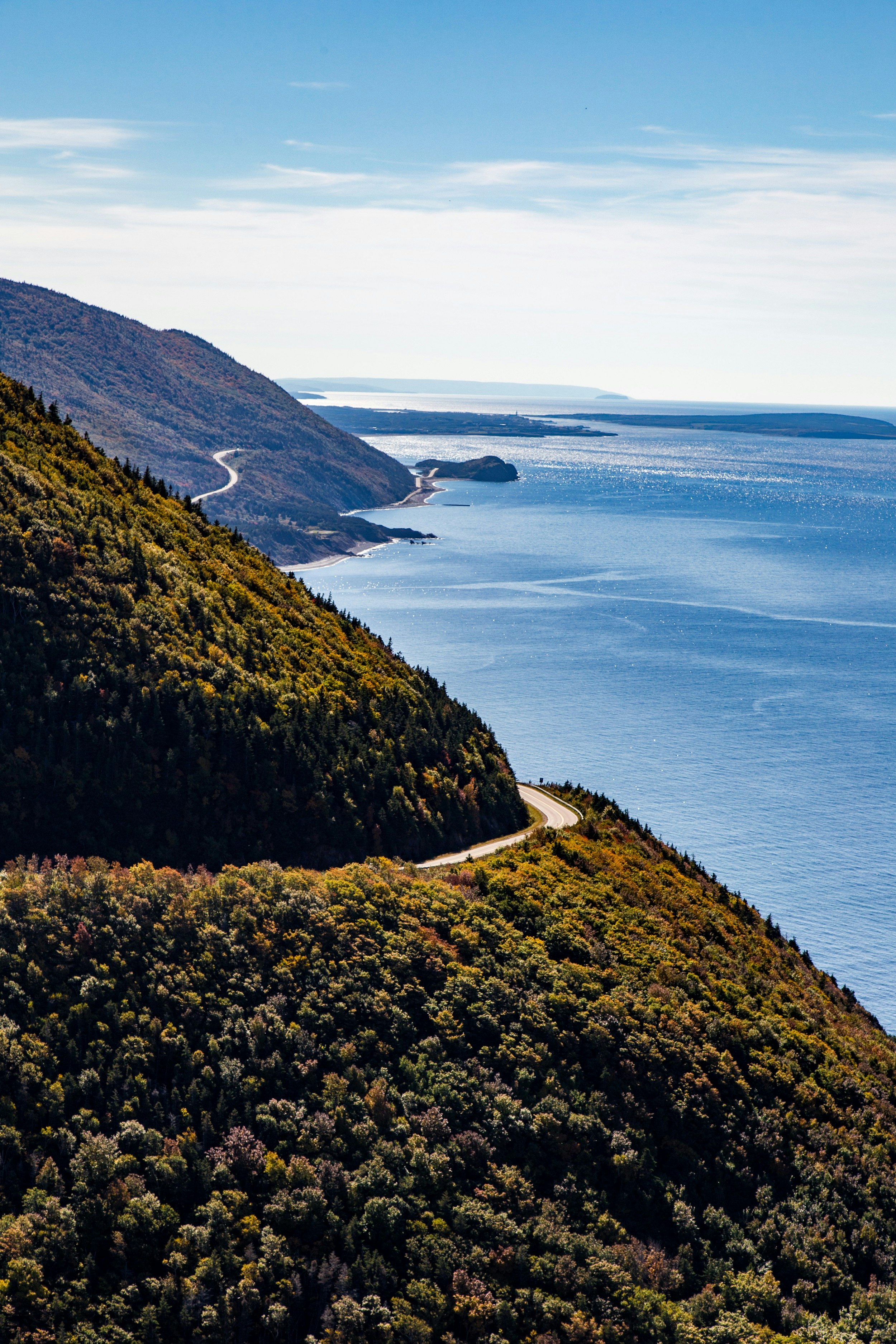 This image looks along a coastline, which has two forested promontories sticking out into the ocean; cut into both is the snaking Cabot Trail. 