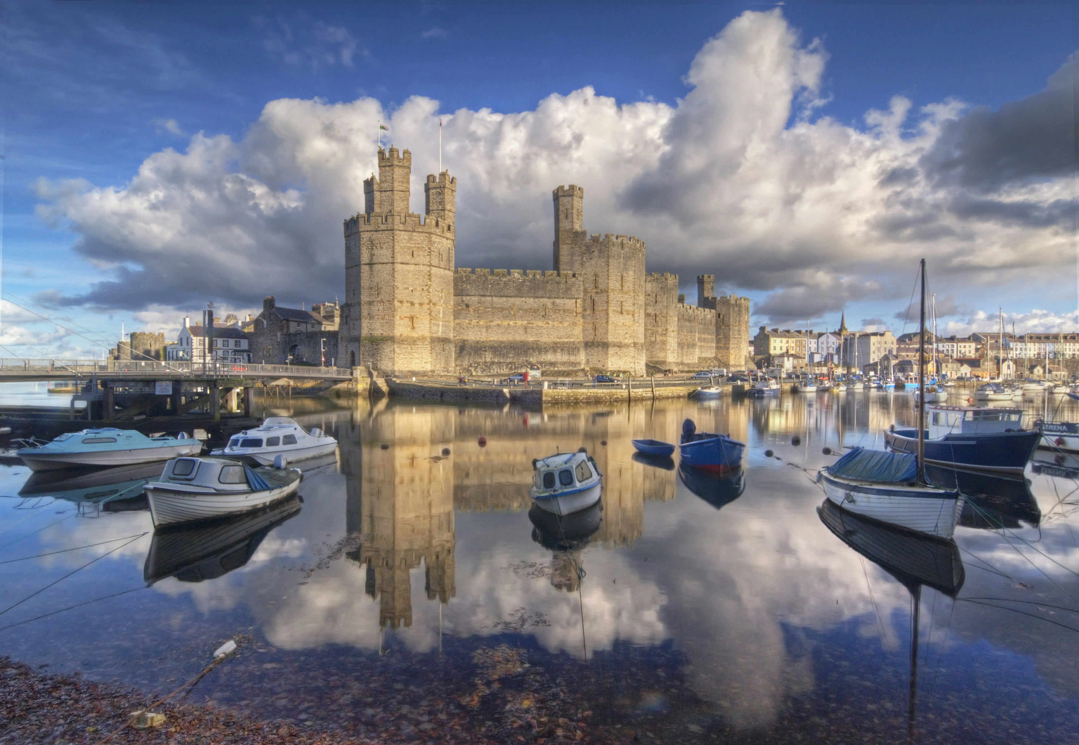 Several small boats sit on the glassy water surrounding Caernarfon Castle in Wales; picturesque waterfront buildings can be seen in the background.