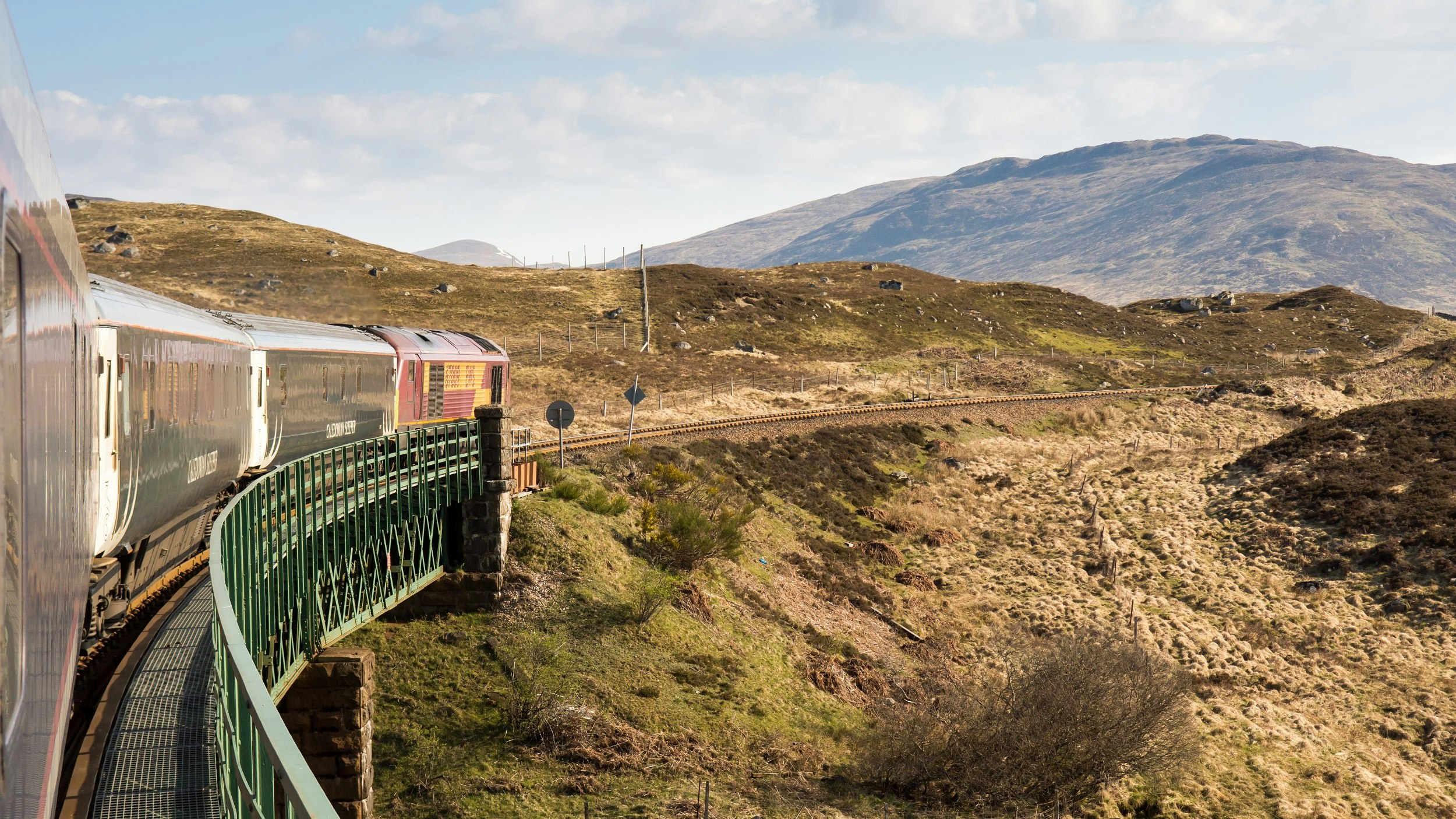 The Caledonian Sleeper train crosses Rannoch Viaduct on the scenic West Highland Line railway in the Scottish Highlands; the image is taken from the train on a curve, so you see the train arching ahead through a barren looking section of rolling hills..