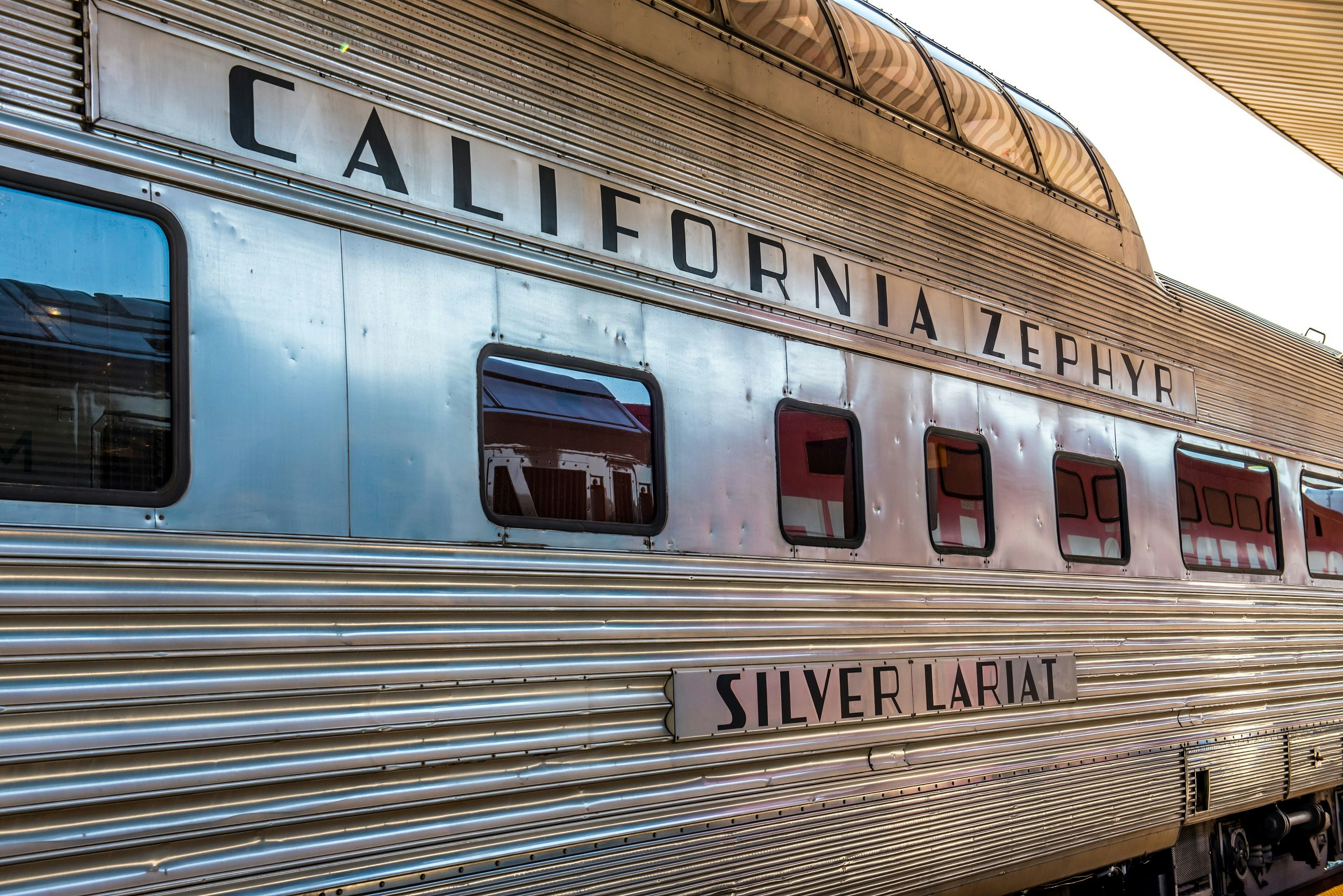 A silver train car with a glass rooftop viewing area sits at a station platform; emblazoned on the side is California Zephyr and Silver Lariat.