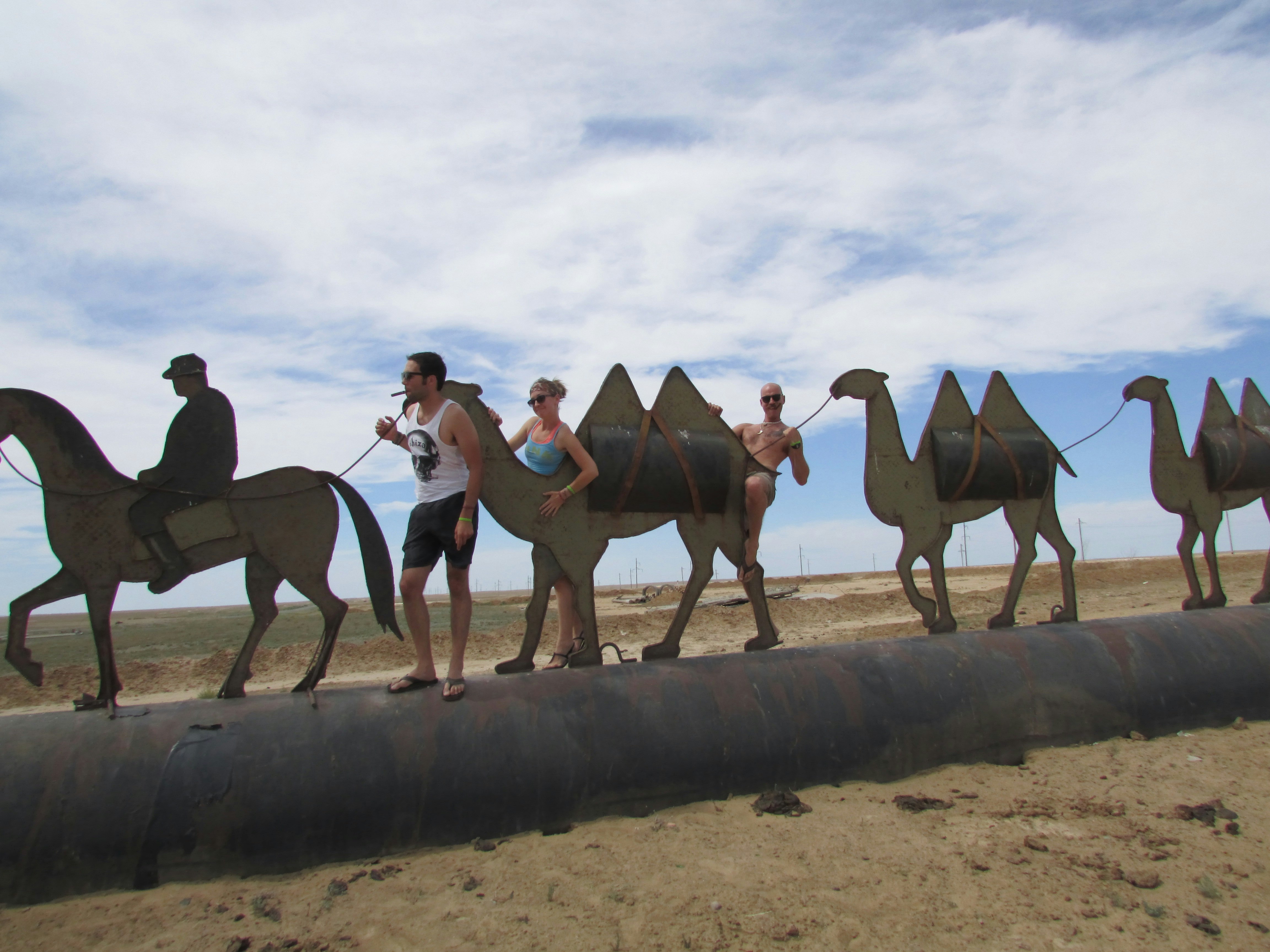 Mongol Rally participants pose with a thin metal sculpture of a camel train, standing behind and beside it to look like they're riding the camels.