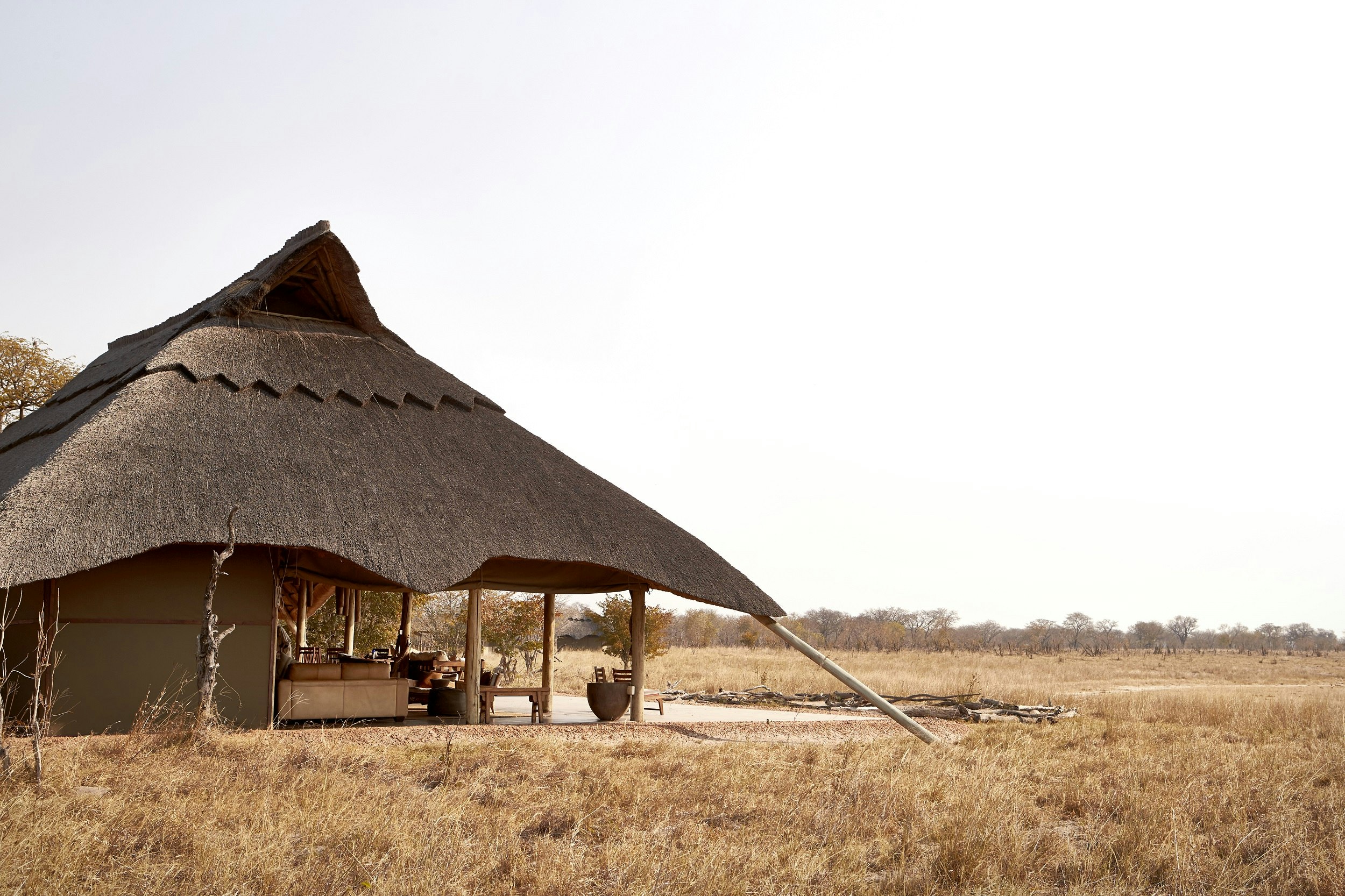 A beautiful thatch-roofed safari accommodation lodge sitting in a dry grass savannah; one half of the interior is open to the environment, with just the roof covering it (no walls).
