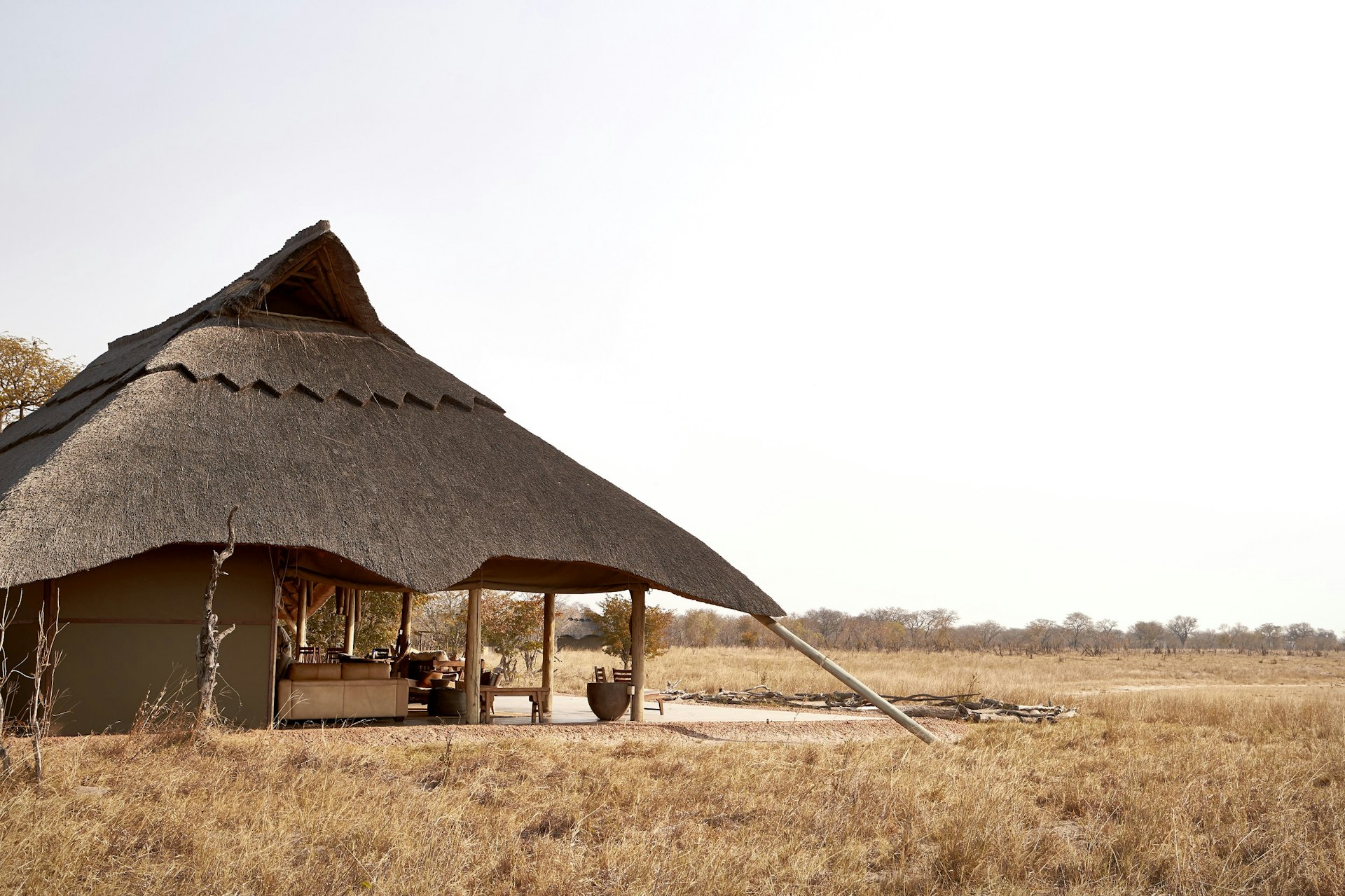 A beautiful thatch-roofed safari accommodation lodge sitting in a dry grass savannah; one half of the interior is open to the environment, with just the roof covering it (no walls).
