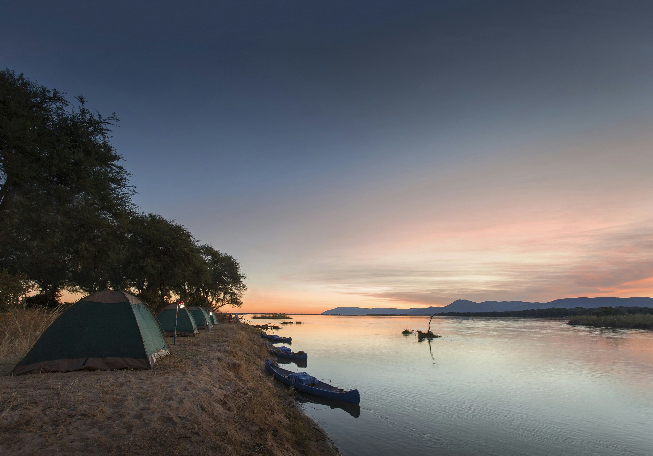 A group of tents sits in a line along the edge of the Zambezi river at sunset; trees flank the tents and canoes are moored on the steep bank.