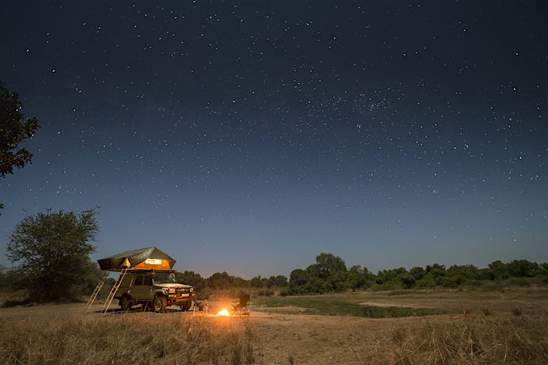 A 4WD with tent raised atop it (with two ladders down to the ground) sits on a grassy plain next to a tree and under a star-filled sky; in front of the truck are two folding camping chairs and a roaring campfire.