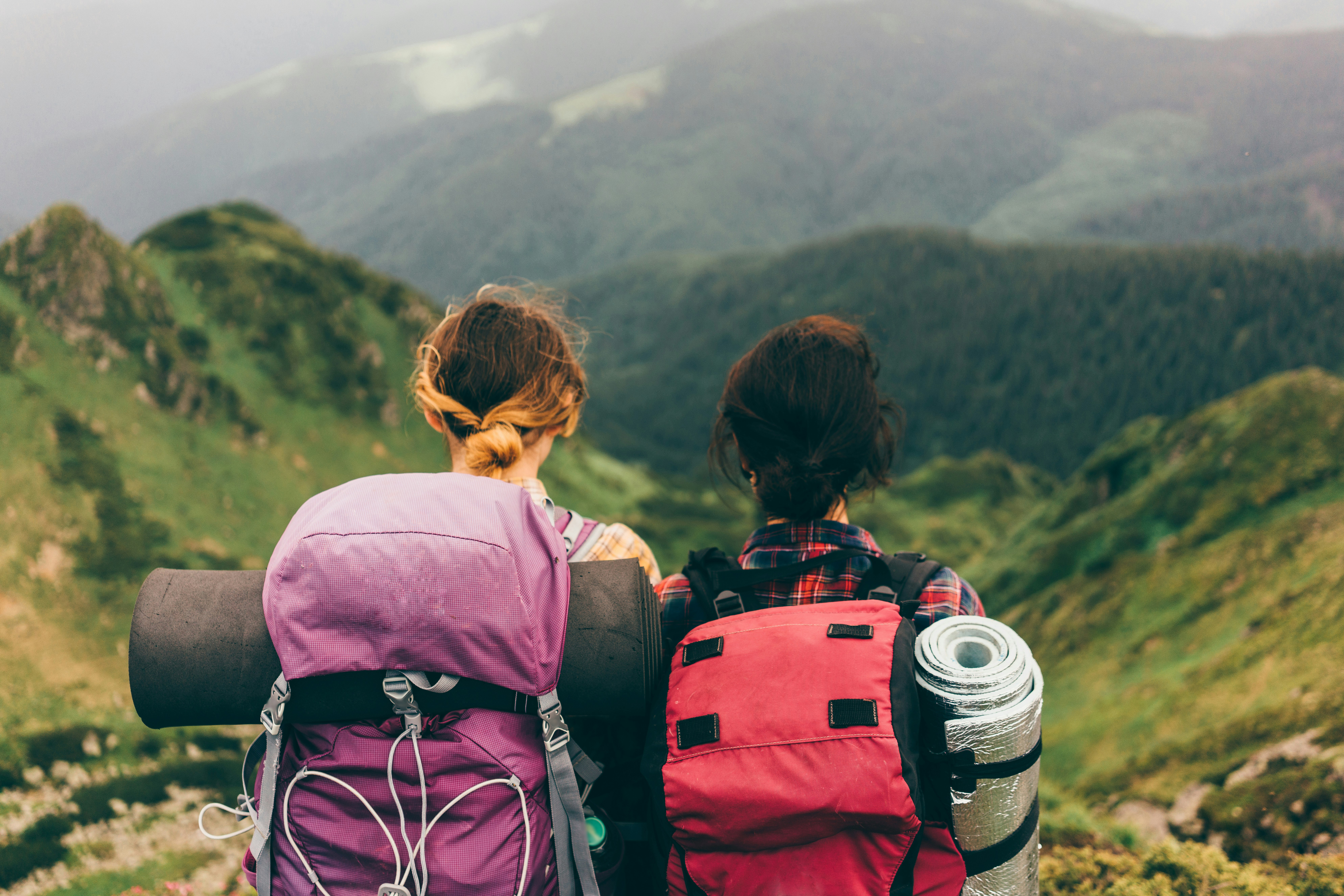 Two women stand facing rolling green hills while wearing backpacks full of camping gear.