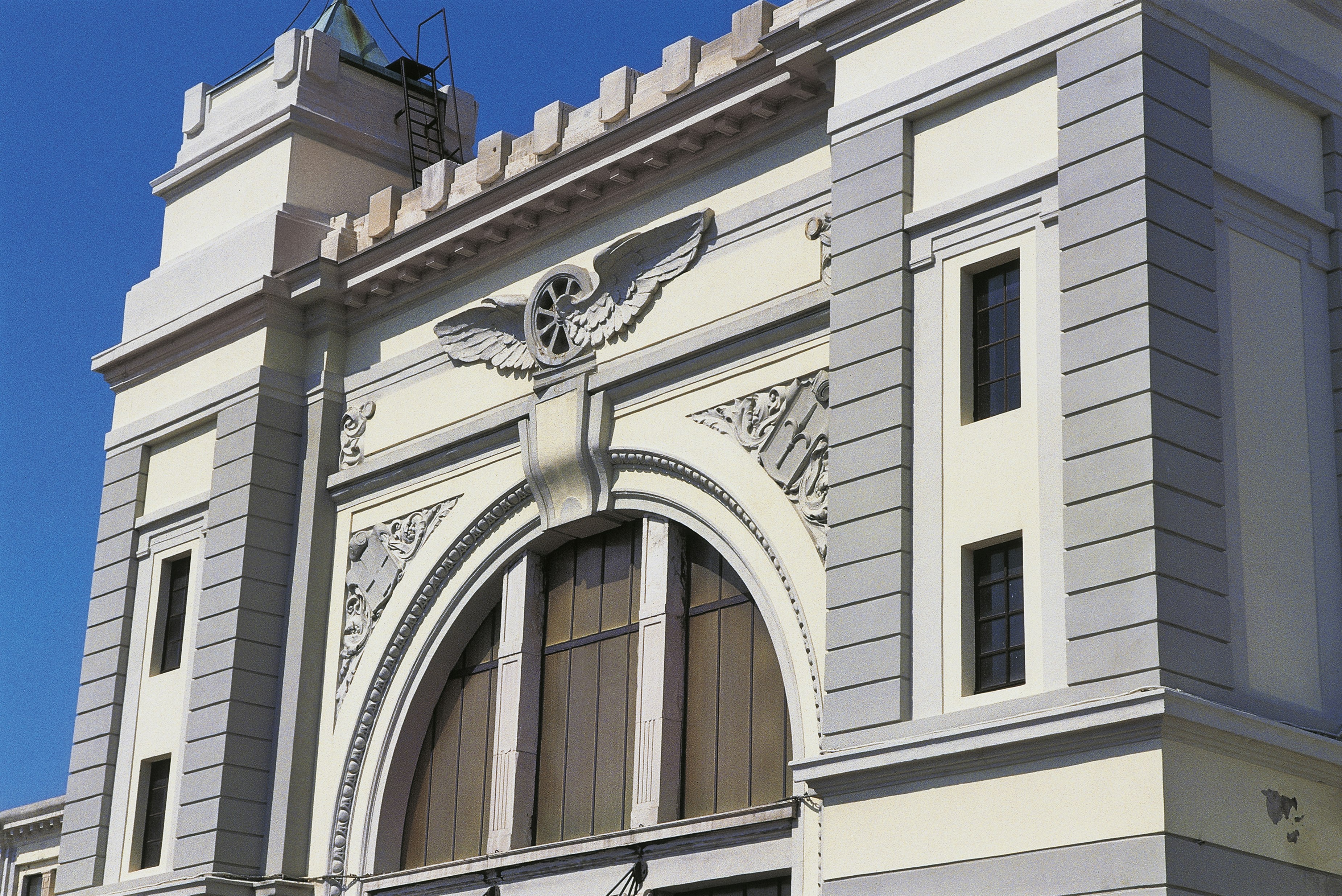 The white and grey stone facade of the former Campo Marzio train station in Treiste is made of two large square columns on either side of a grand arch-shaped window. Each column has two narrow rectangular windows, while the windows that once filled the arch have been filled in. Over the arch is a wheel with wings carved into the stone. Behind the structure is a brilliant clear blue sky.