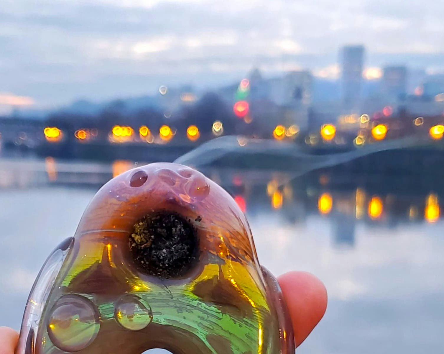 A rainbow-hued pipe packed with cannabis is held up in front of the Portland, Oregon skyline at dusk