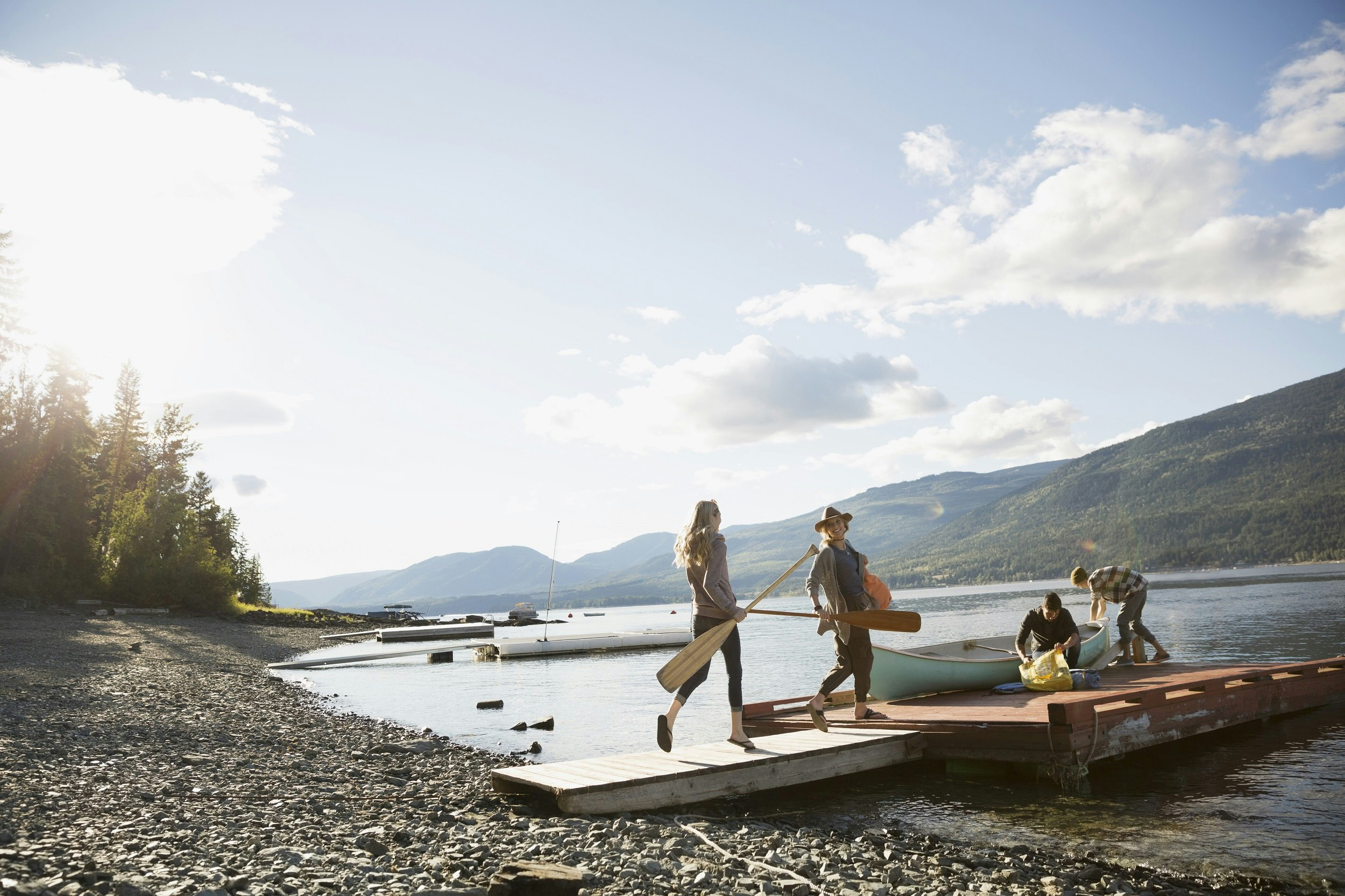 Two people carrying paddles walk onto a dock where two others are loading a canoe with supplies; the dock is off a rocky beach, and the lake is calm, with forested hills in the background.