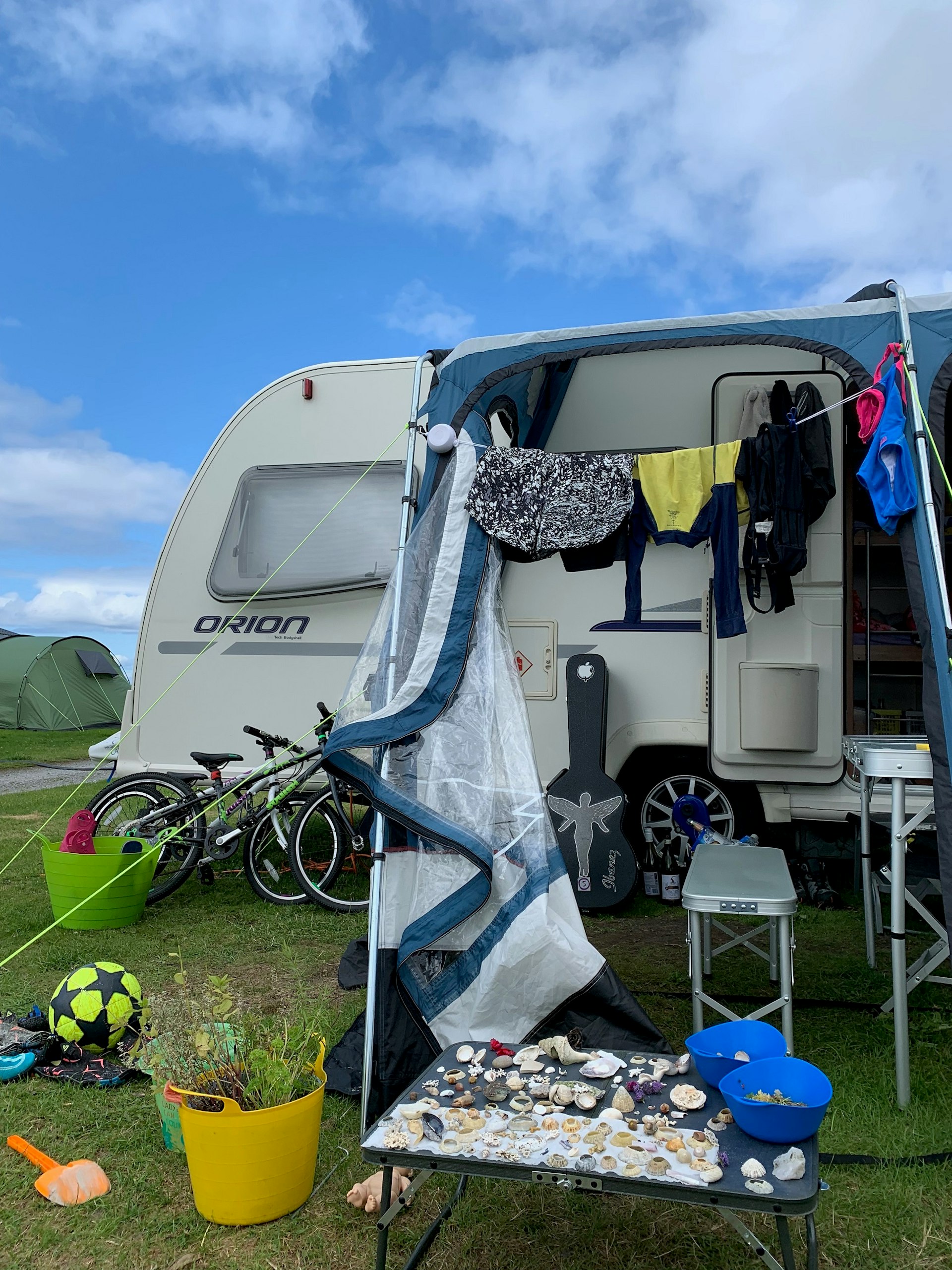 Exterior of a caravan with items scattered outside including bikes, a football, a spade, a guitar, a table covered in seashells and a washing line full of clothes.