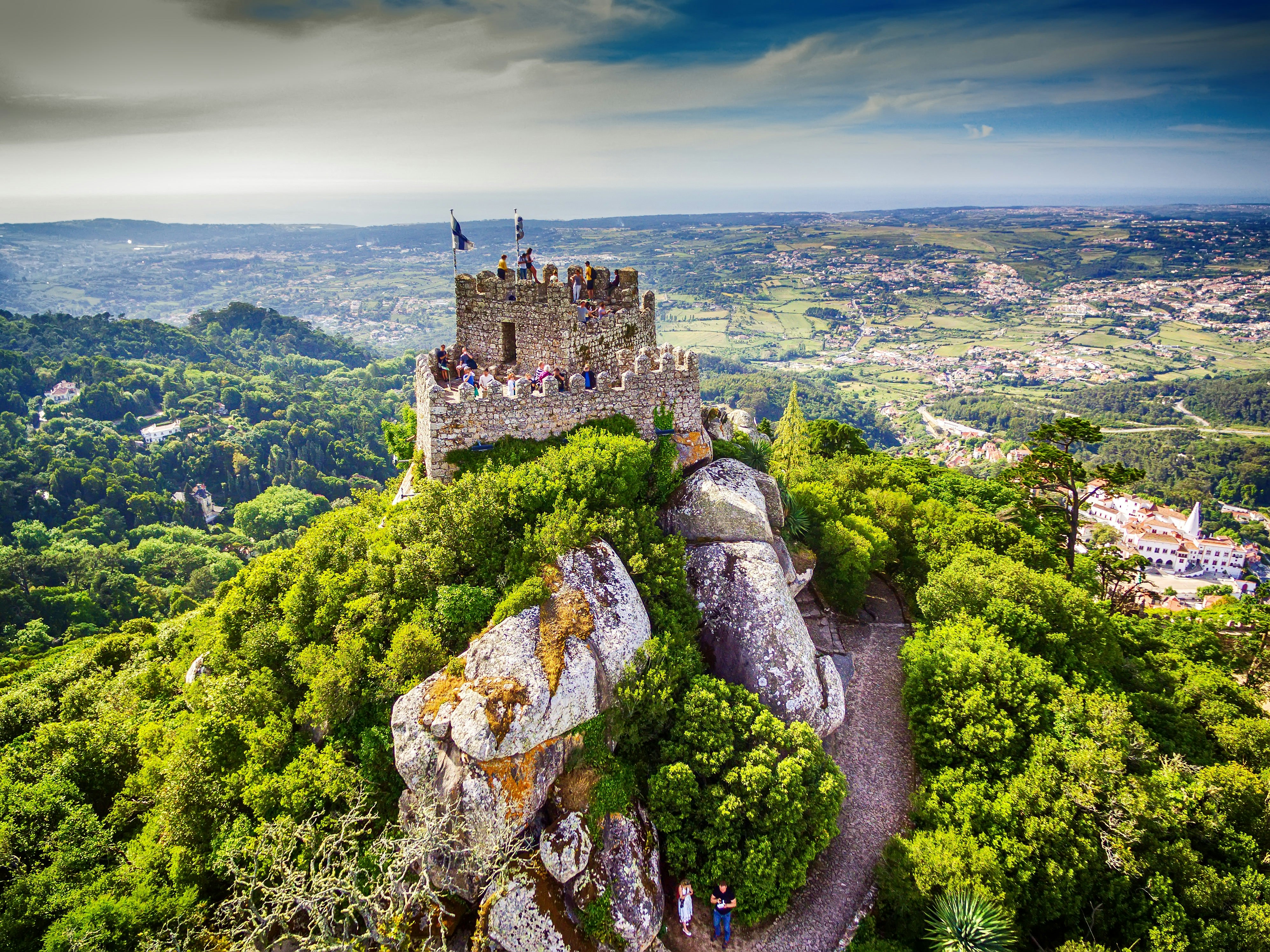 Aerial view of Castelo dos Mouros (Castle of the Moors) in Sintra; the ancient castle sits atop a greenery-covered hillside, and people are standing in the tower, looking out over the valley below.
