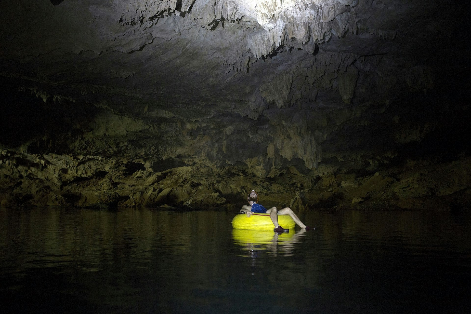 A lone person wearing a head torch sits in a yellow inflatable tube and floats through the waters in the darkness of a cave.