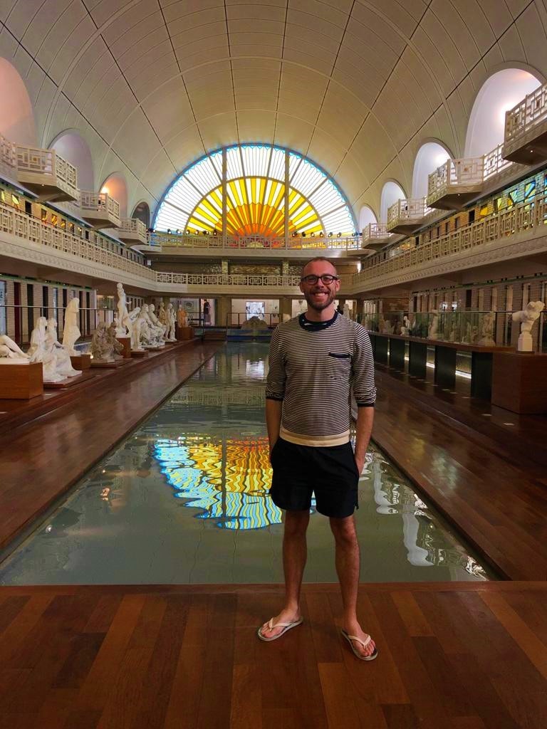 Writer Tom stands in front of an Olympic sized art deco swimming pool lined with sculptures; at the end of the room is a stained glass window that resembles the rising sun.