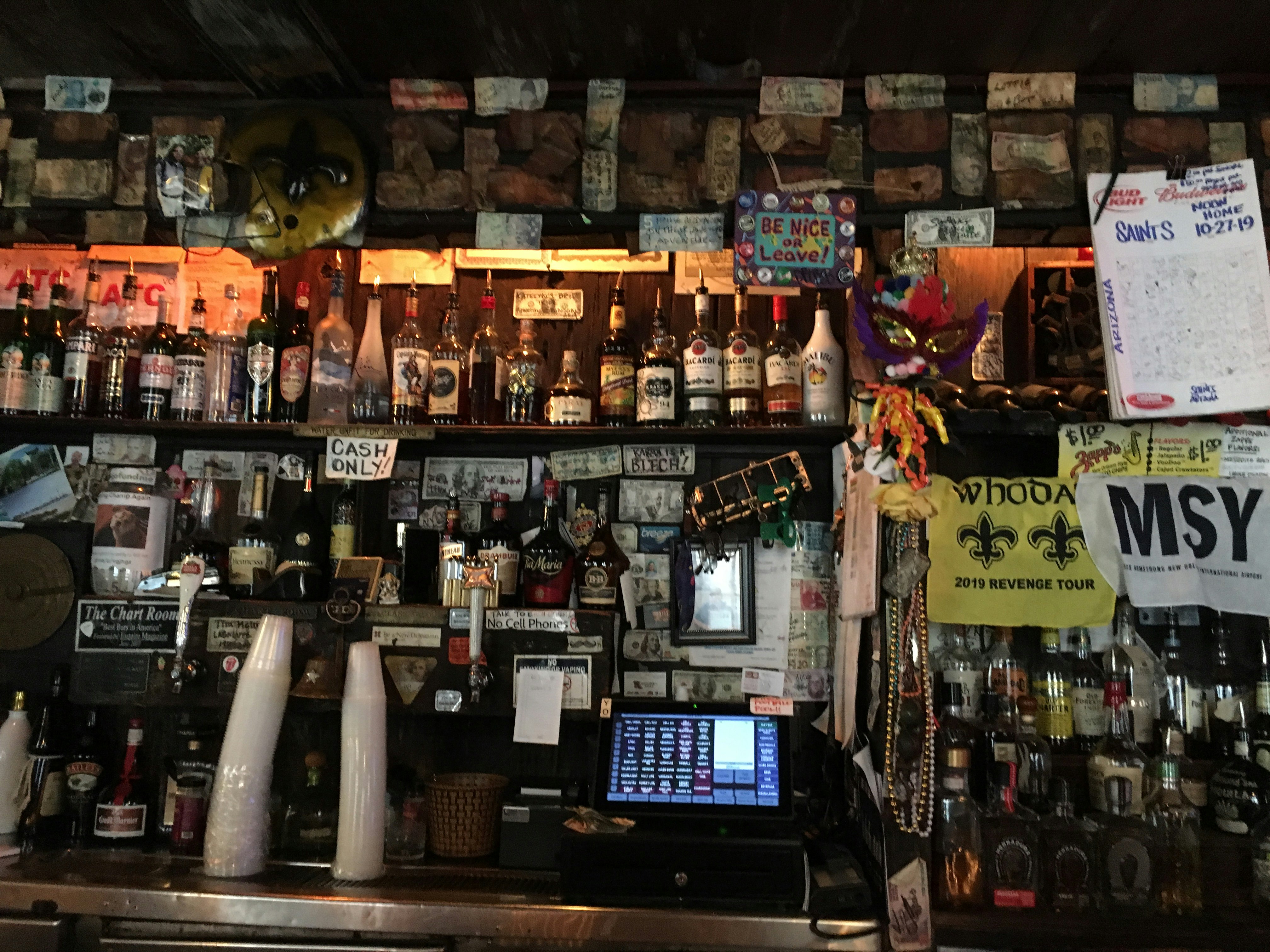 View behind the bar at the Chart Room. Shelved are filled with multiple bottles of alcohol and covered in different currencies, Mardi Gras masks and beads. A cash register is off to the left of a stack of plastic cups.  