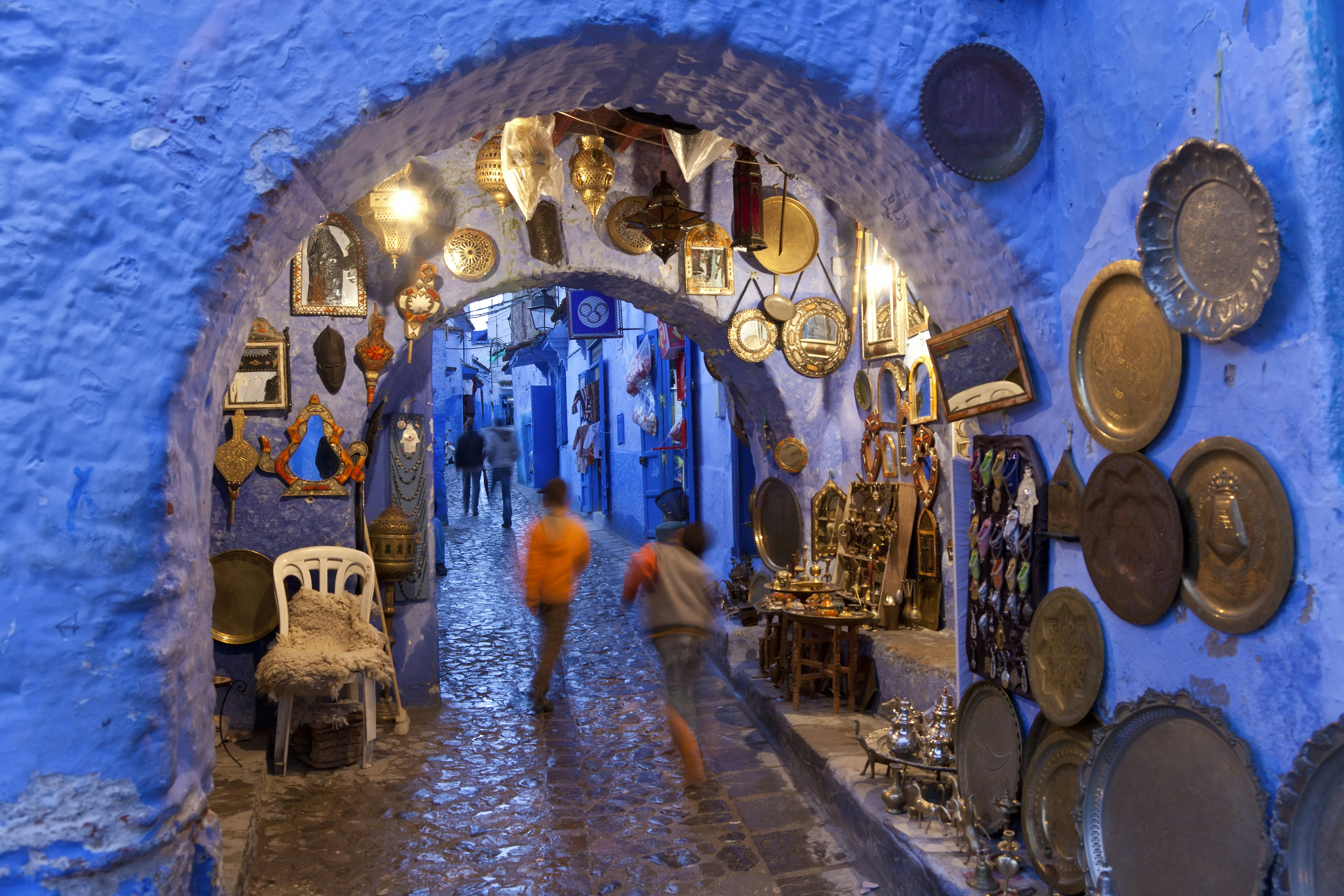 Two children in orange and grey clothes are blurred as the spring through the cobblestone streets of Chefchaouen, surrounded by the blue plaster walls of the medina and shining metal serving treys, mirrors, and other craft goods
