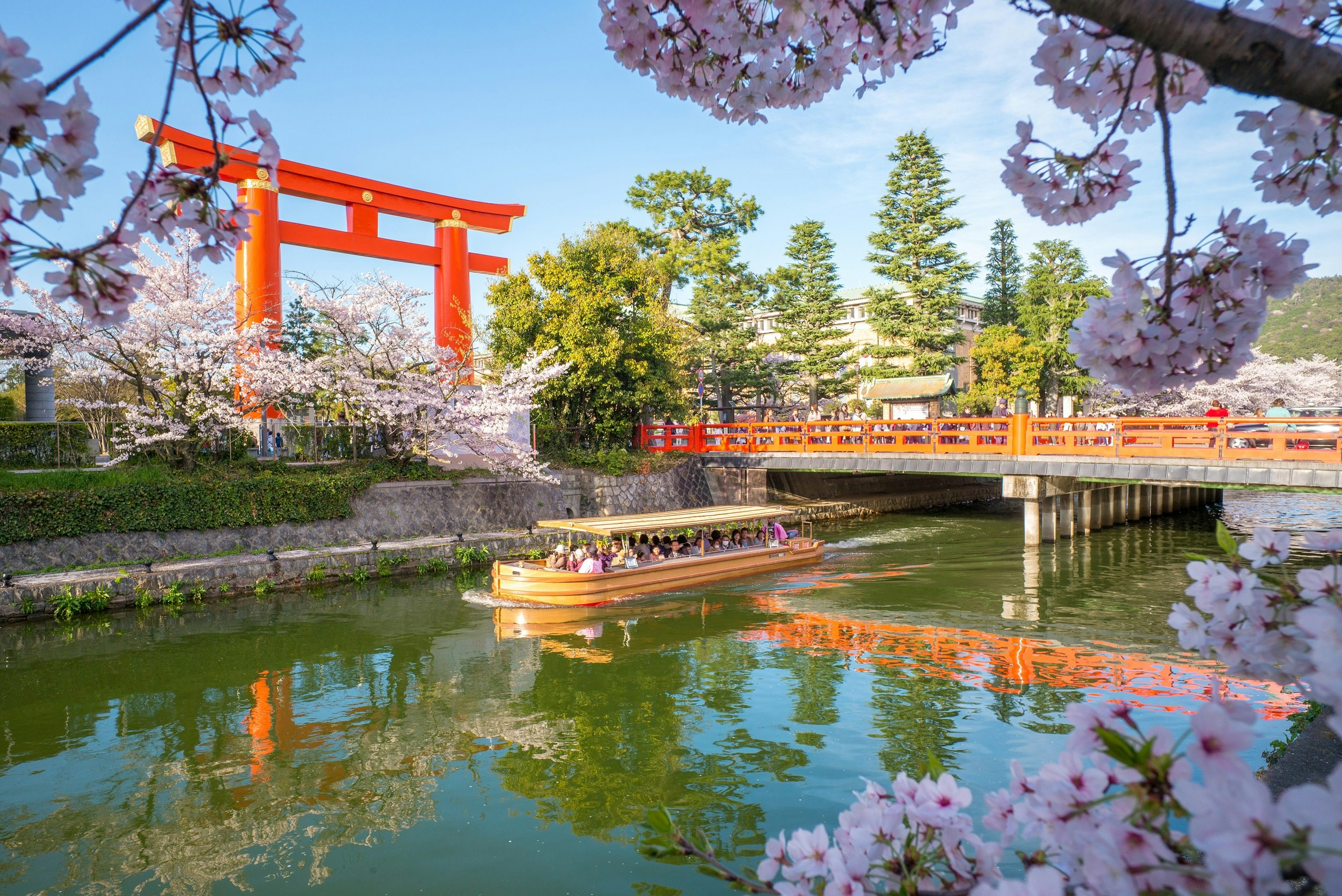 A tour boat sails down the canal past the Heian Shrine lined with cherry blossom and other trees in Kyoto on a sunny day
