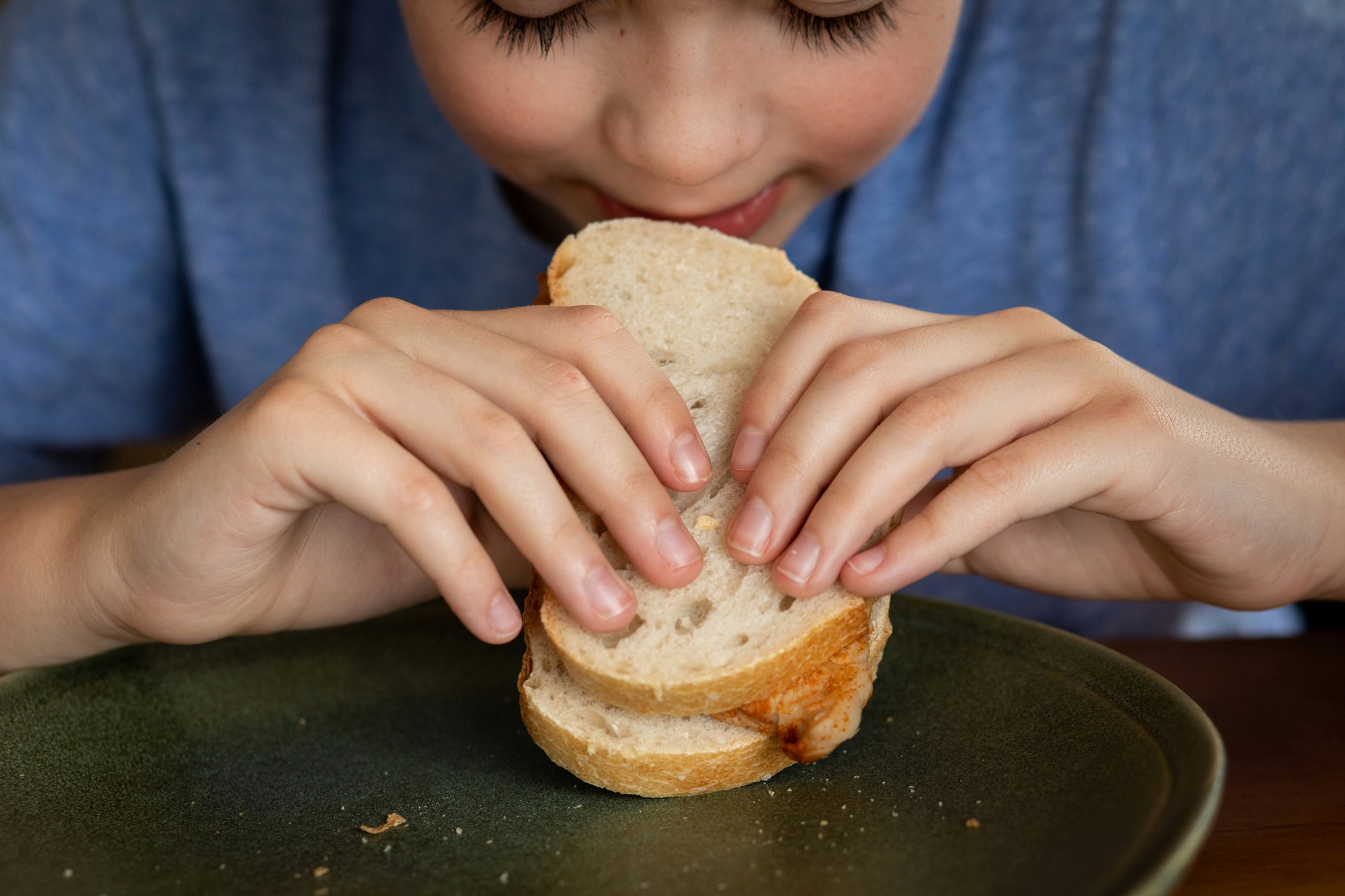 A close-up of a young boy leaning in to take a bite of a bifana sandwich; marinated pork between two slices of bread.