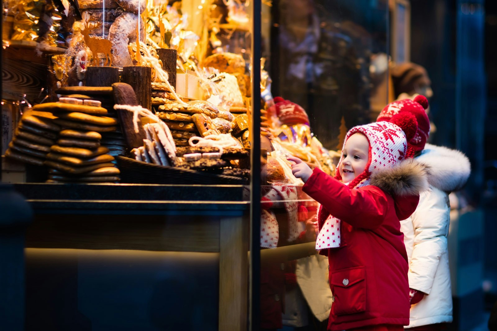 A smiling toddler points to an illuminated display of cakes and pastries at a Christmas market