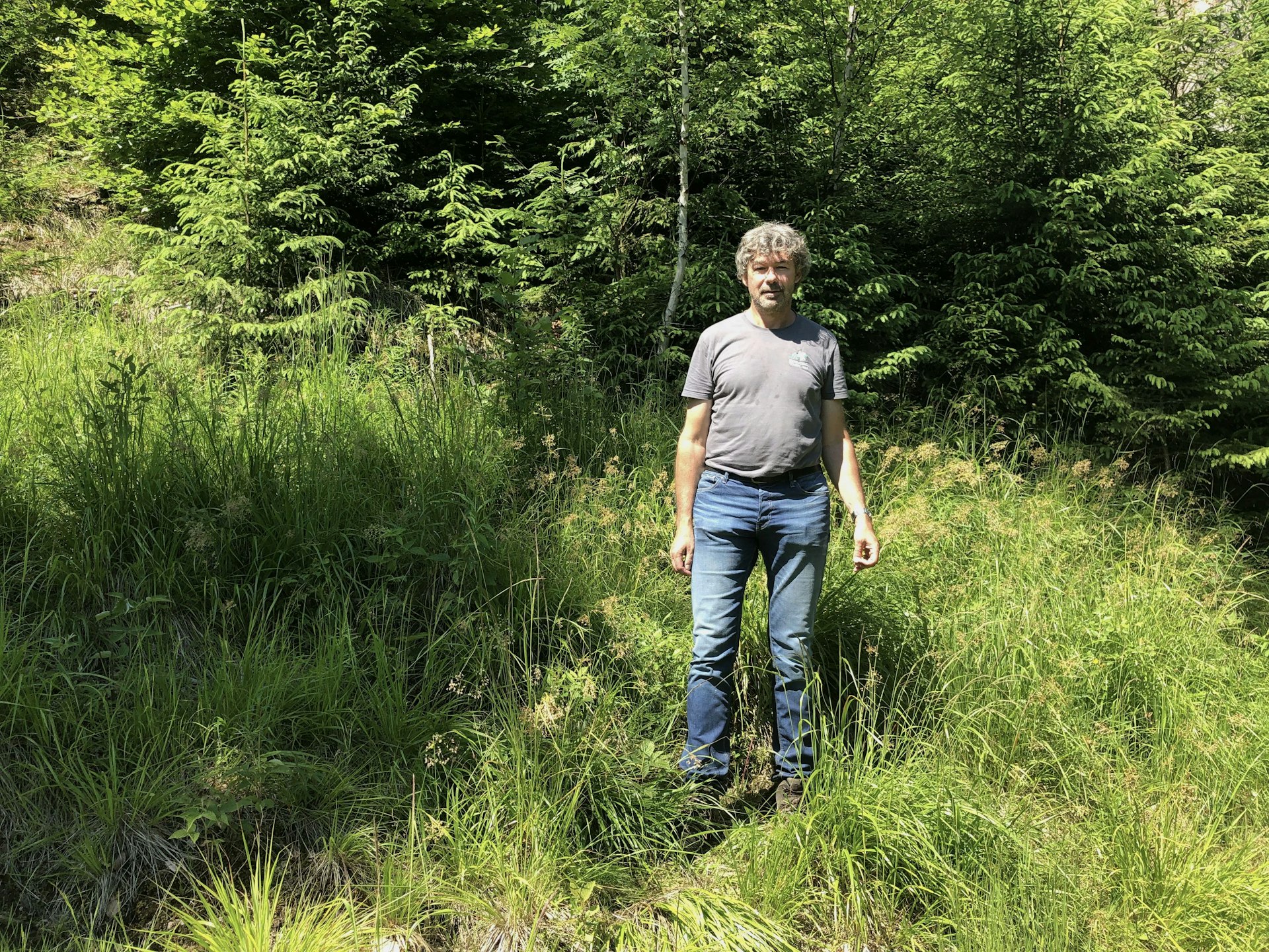 Chistoph Promberger stands against a forest backdrop wearing jeans and a t-shirt. His hair is grey.