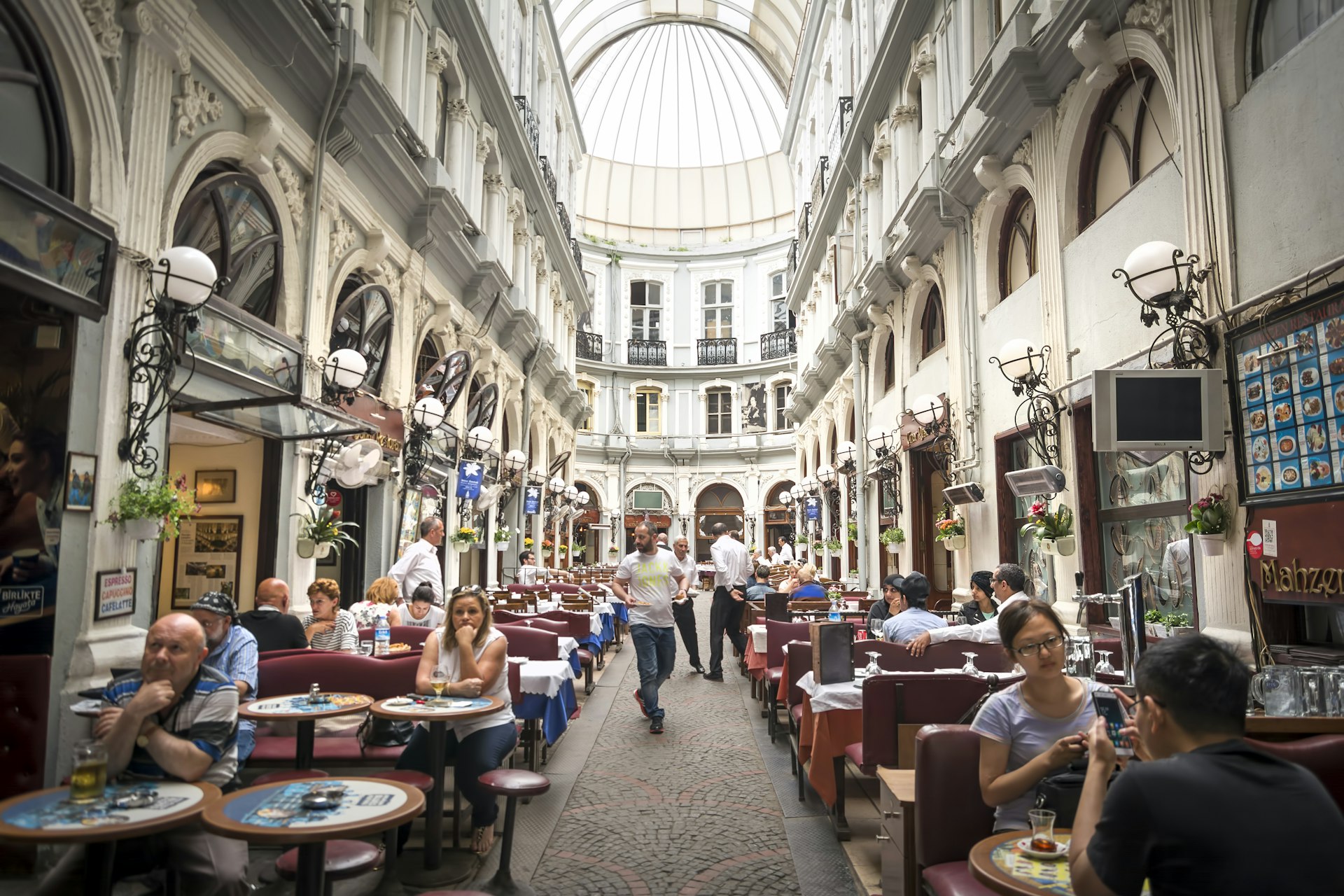 Many people are sitting at the tables of the cafes which line an arcade in Istanbul; the facades are ornately decorated with carvings and lamps.