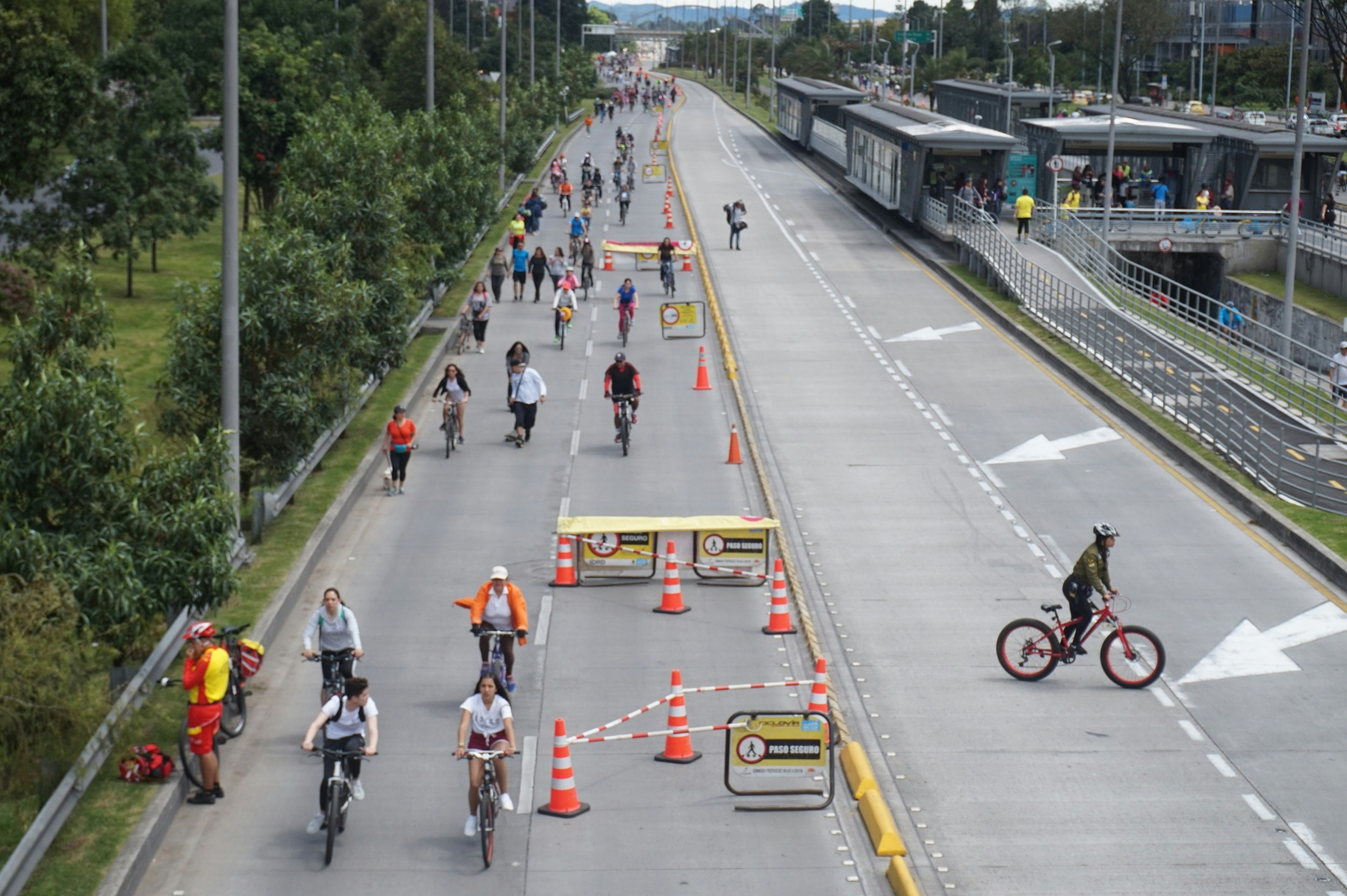 Looking down a main road from above, two lanes are filled with bicycles riding toward the camera; the opposite side of the road is empty bar a police person on a bike. A paramedic is standing next to their bike by the side of the road.