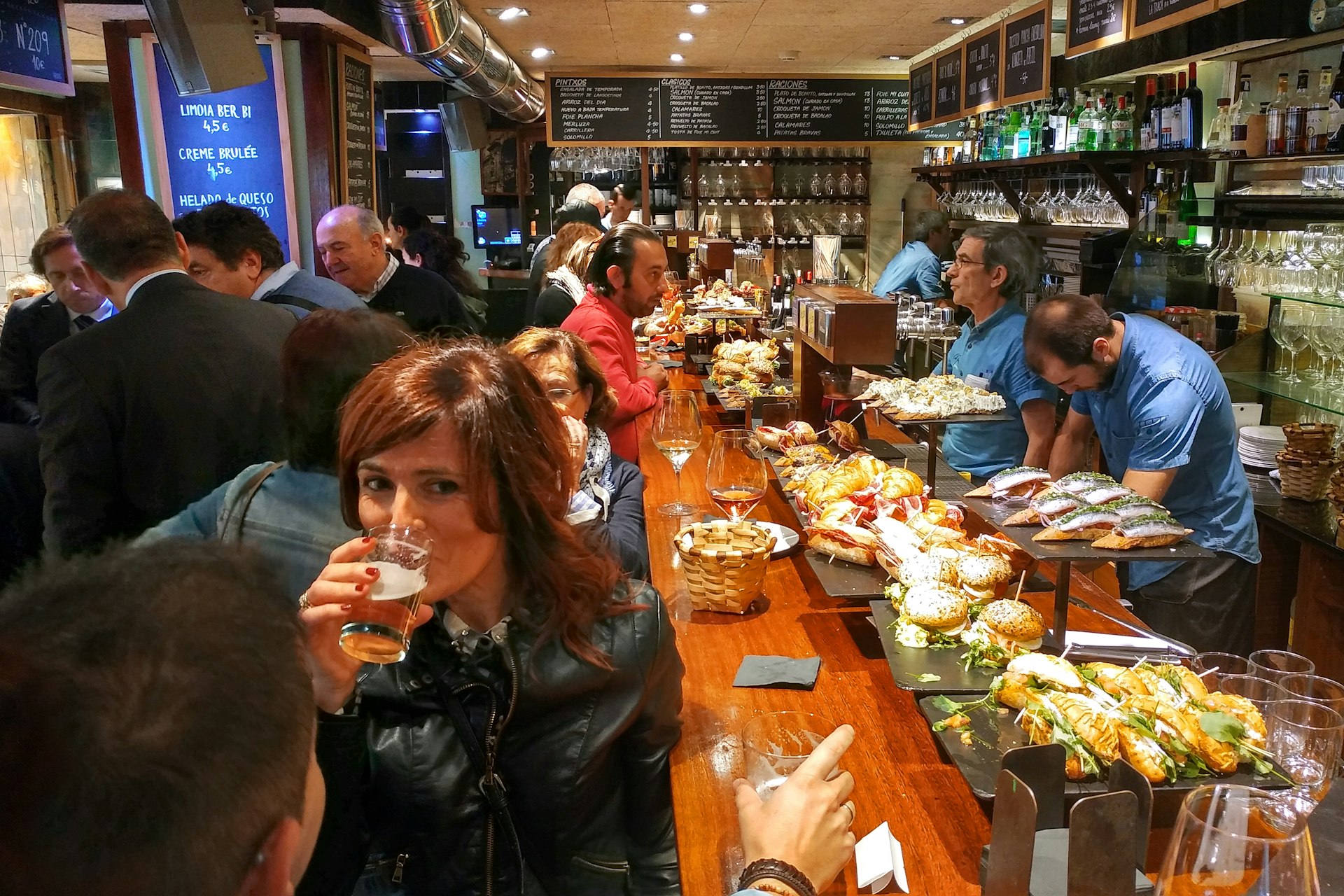 A crowd of people packed into a small restaurant sip ciders and wine set on a long wooden bar. Behind the bar are pintxos on tall metal and wooden stands. Three employees in blue shirts prepare food and beverages.