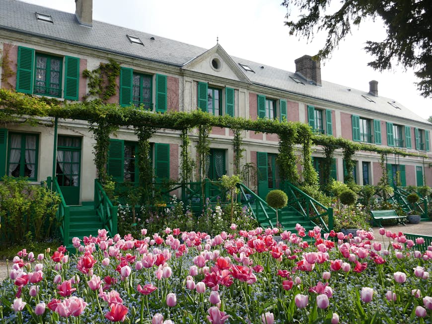 Дом и сад Клода Моне;  большой дом выкрашен в розовый цвет и имеет зеленые ставни.  Возле дома цветет масса розовых цветов.