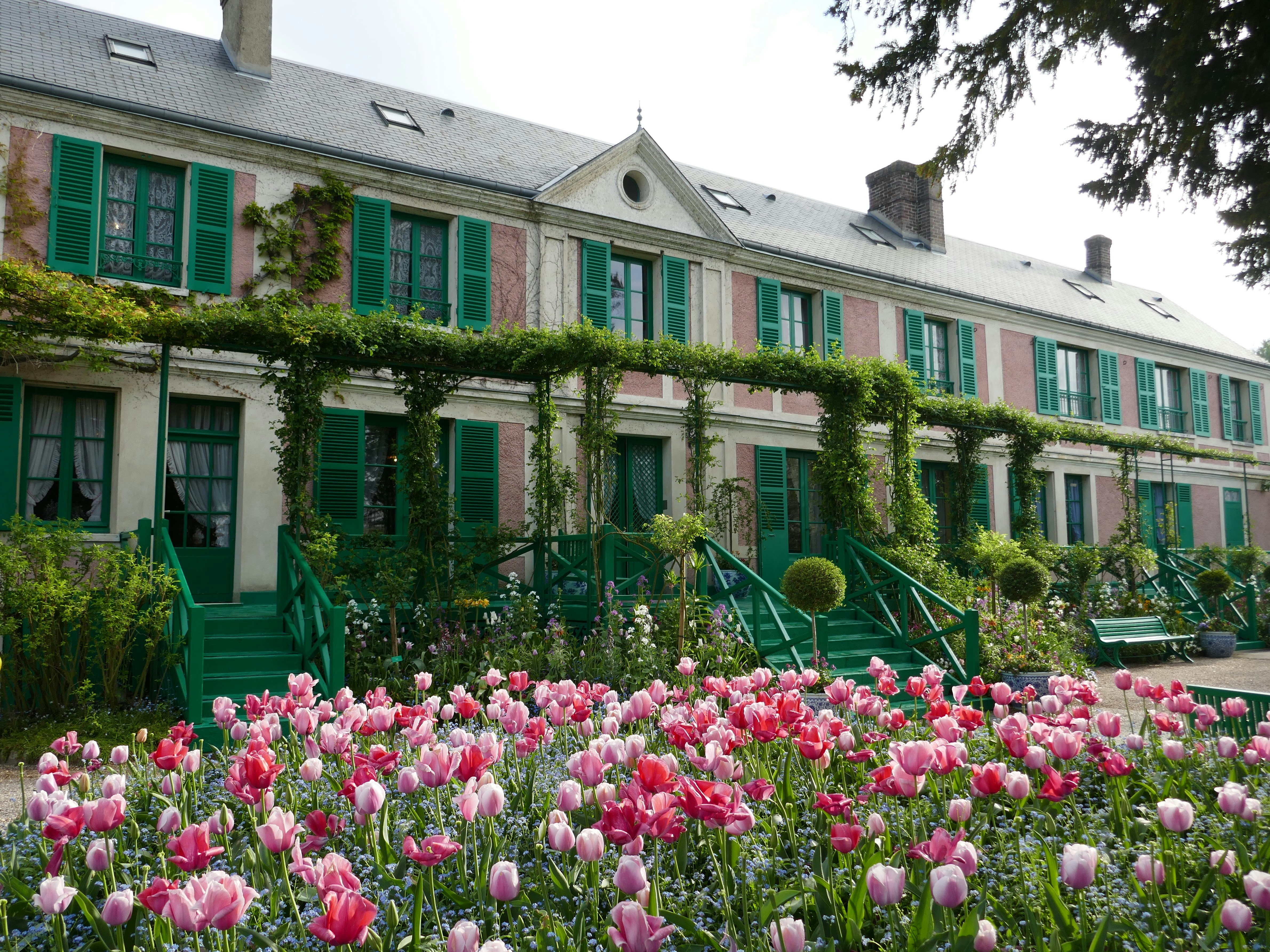 The Maison et Jardin de Claude Monet; the large house is painted pink and has with green shutters. A mass of pink flowers are in bloom outside the house.