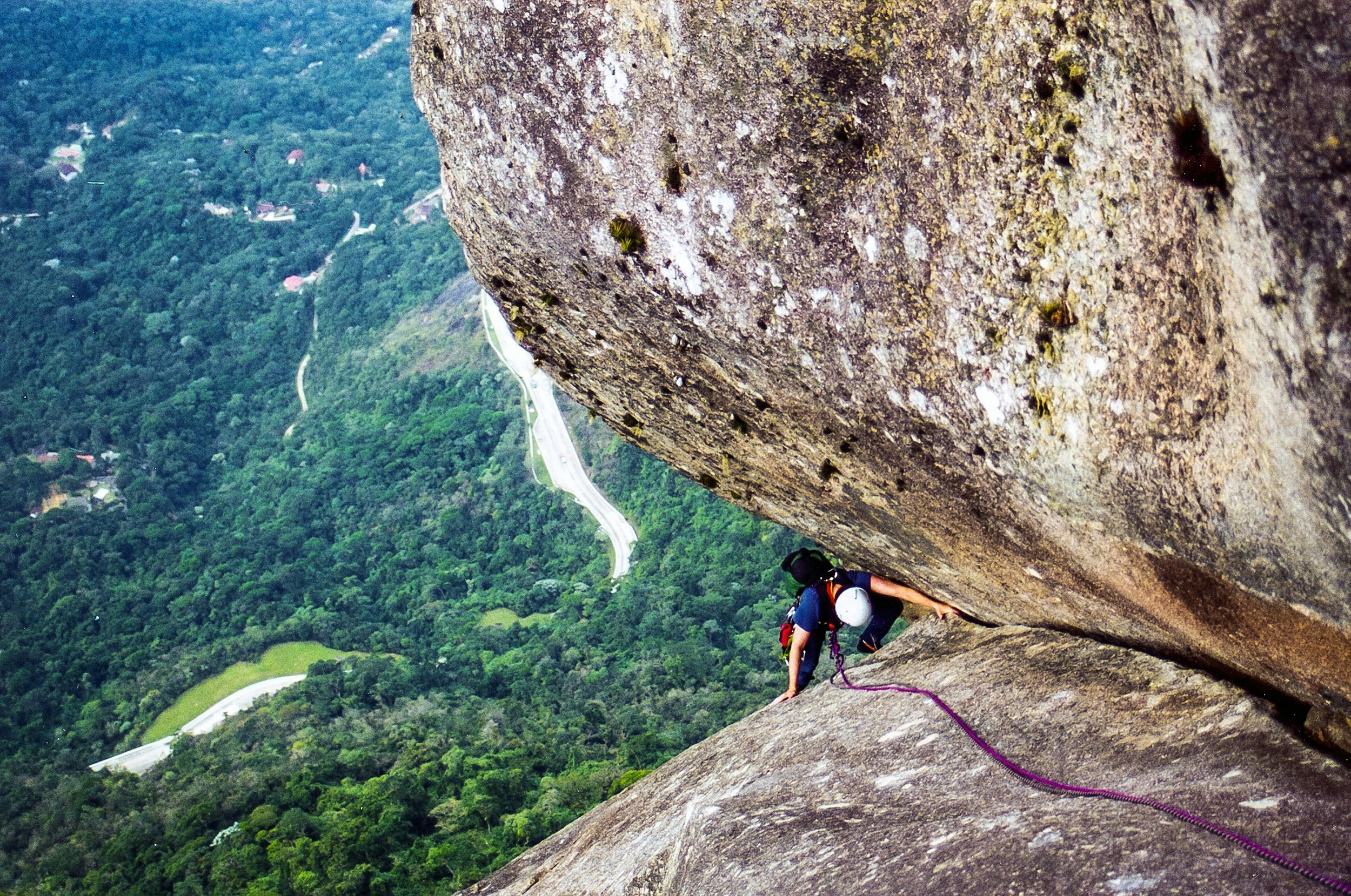 A rock climber ascends a vertical crack between the junction of two massive monoliths; far below in the distance is a forest.