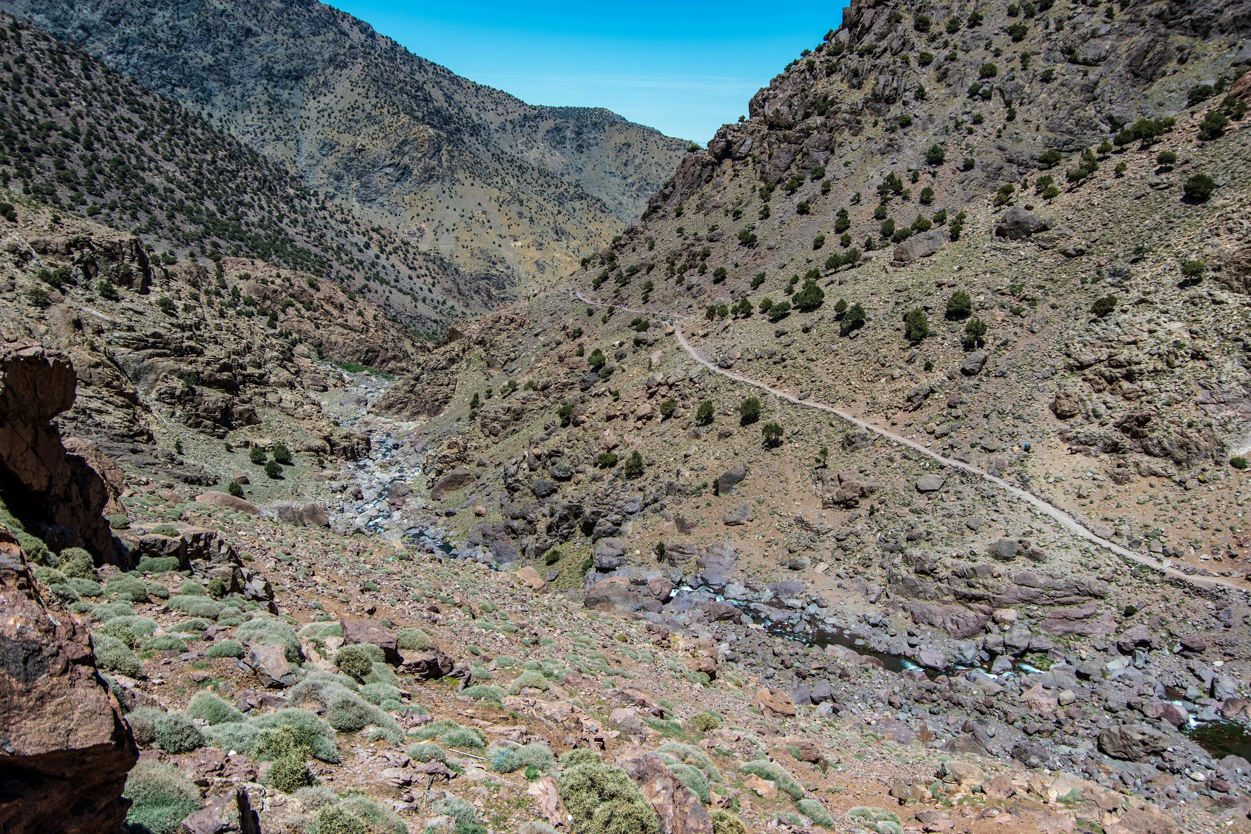 A single track hiking path meanders along the steep side of a rocky valley en route to Jebel Toubkal; the valley is barren, with just some small scrubby bushes dotting the slopes.