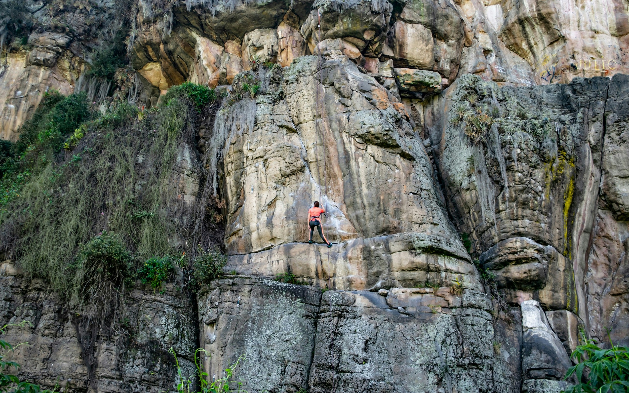 A rock climber stands halfway up a sheer rock slope; they are suspended from a rope above. 