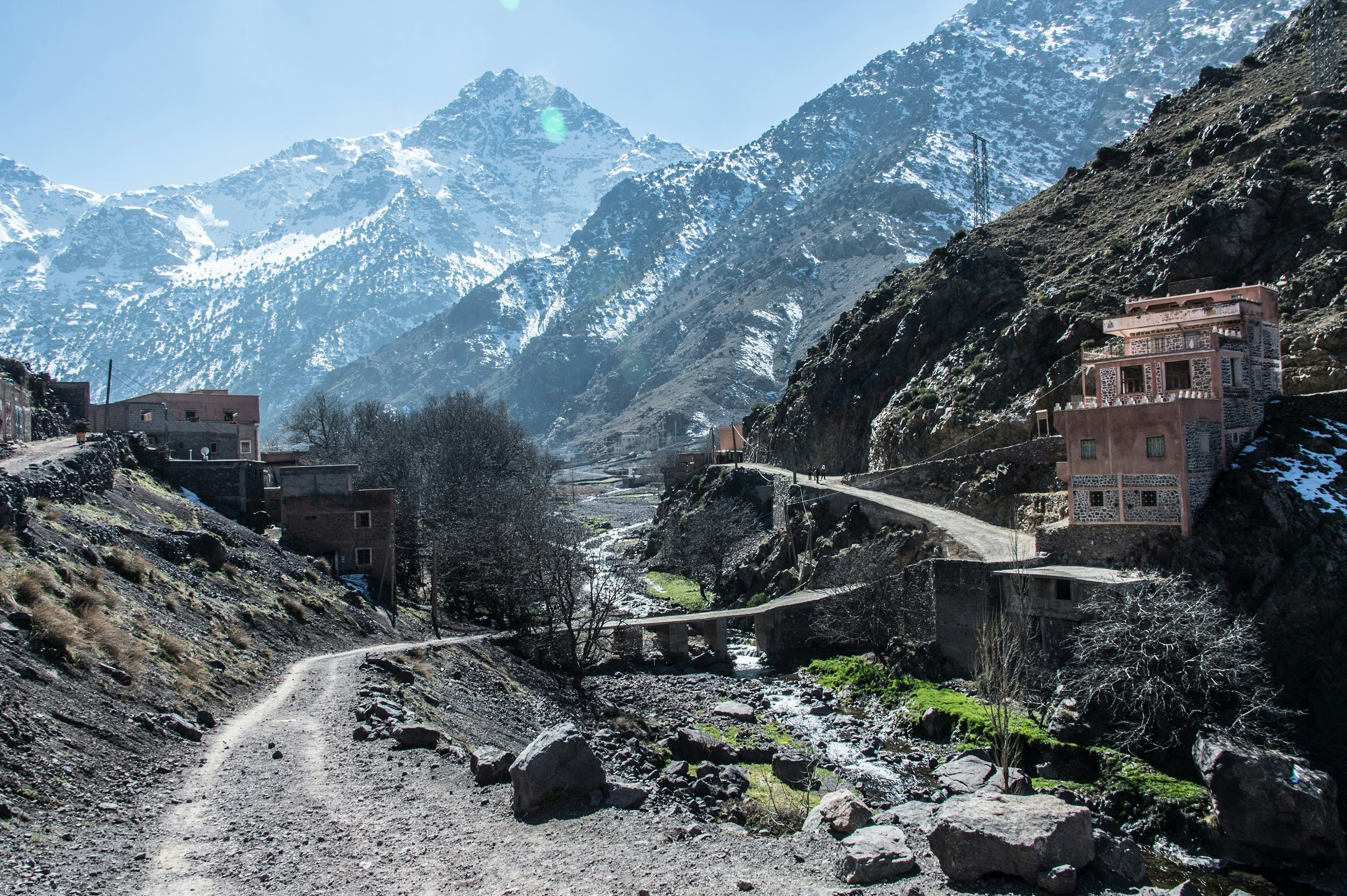 A walking path ascends along the side of a valley towards the distant (and towering) peak of Jebel Toubkal; some small dwellings sit beside the path on the steep hillside.