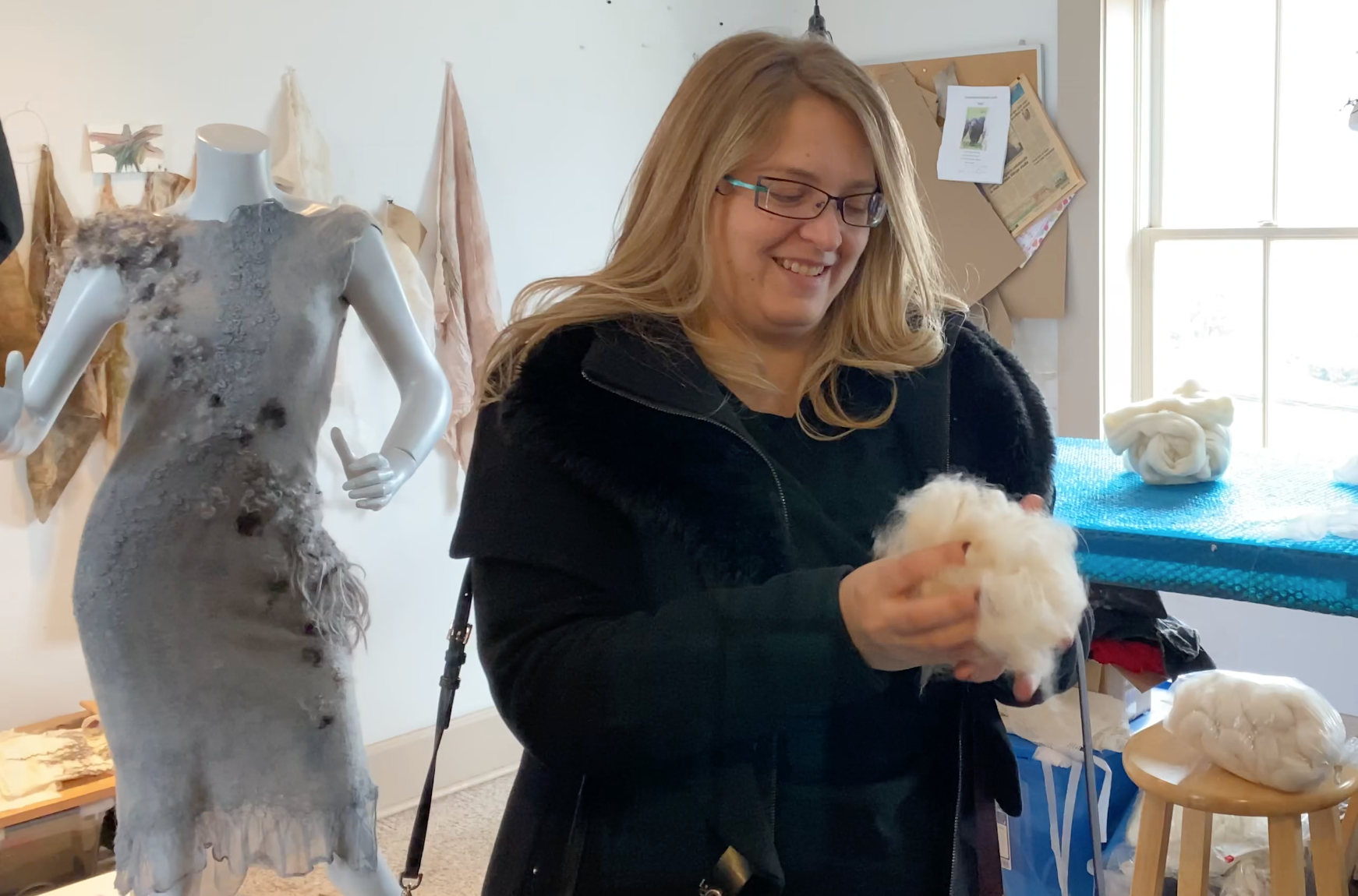 A woman with long blond hair and glasses smiles at a puffball of white alpaca wool. Behind her is a dress by Celeste Malvar Stewart to her right and to her left a long work table covered in blue bubble wrap in front of a bright window