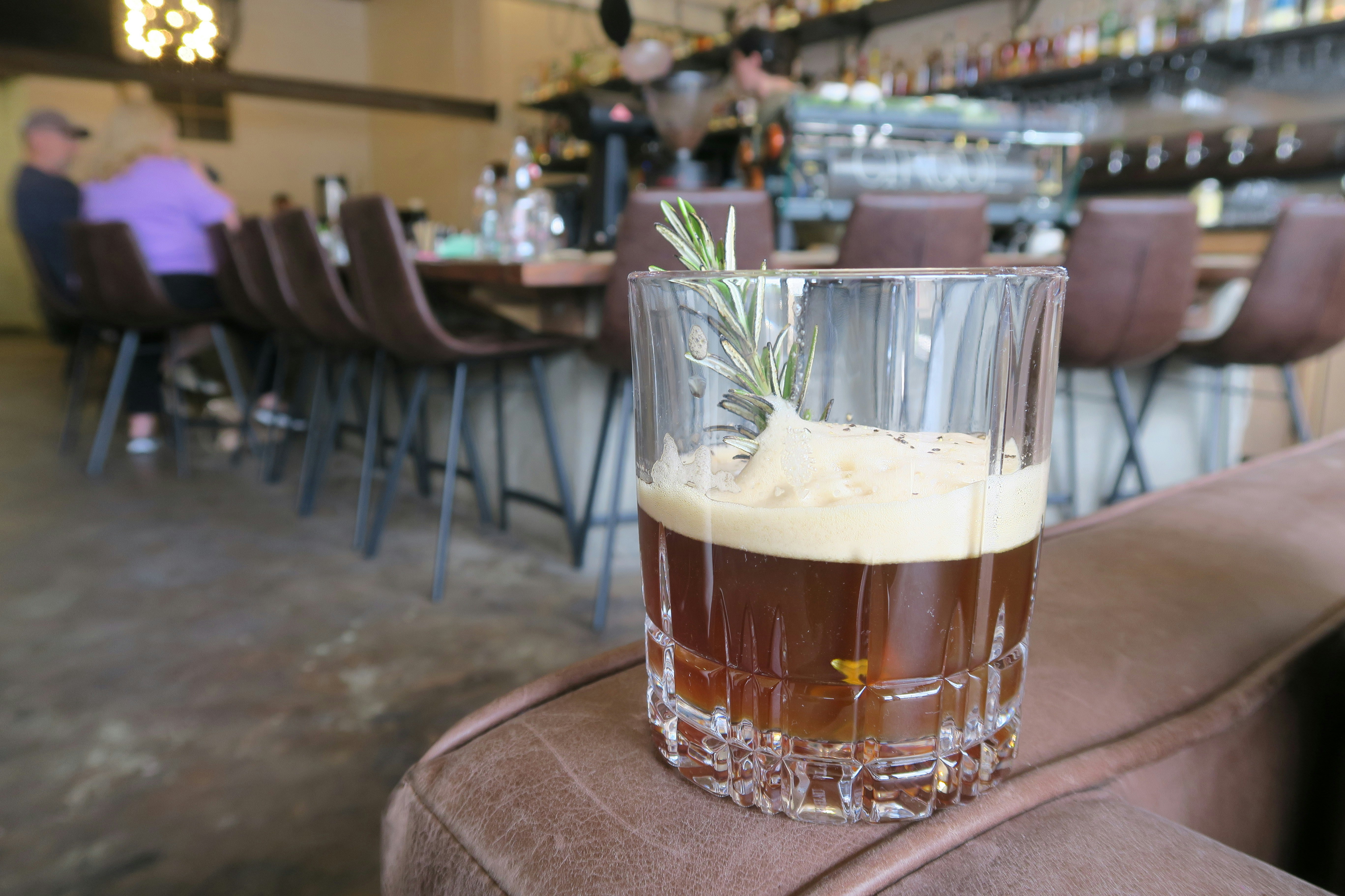 A short cocktail glass filled with a light brown drink garnished with a spring of thyme rest of on an armchair in a bar