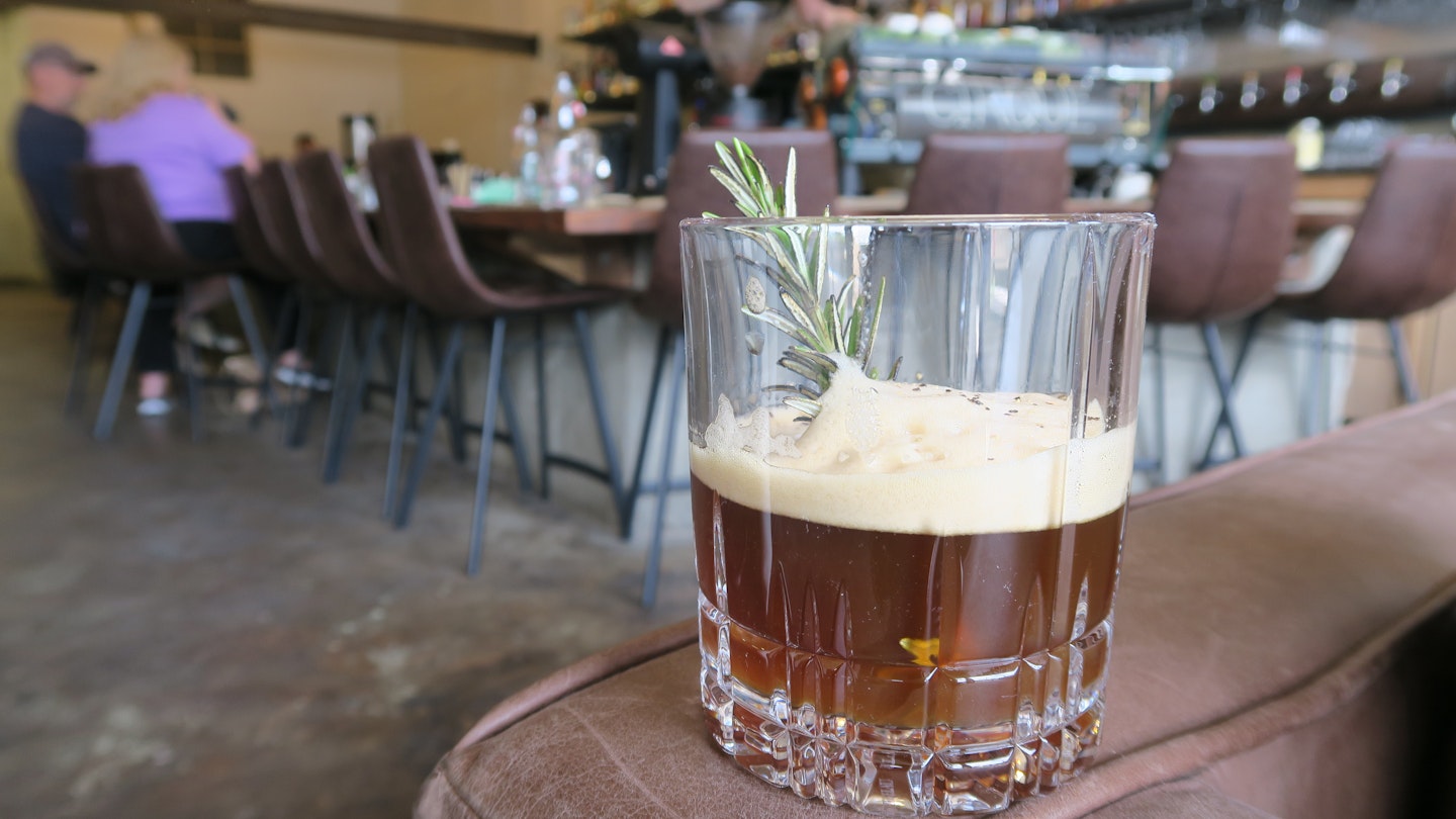 A short cocktail glass filled with a light brown drink garnished with a spring of thyme rest of on an armchair in a bar