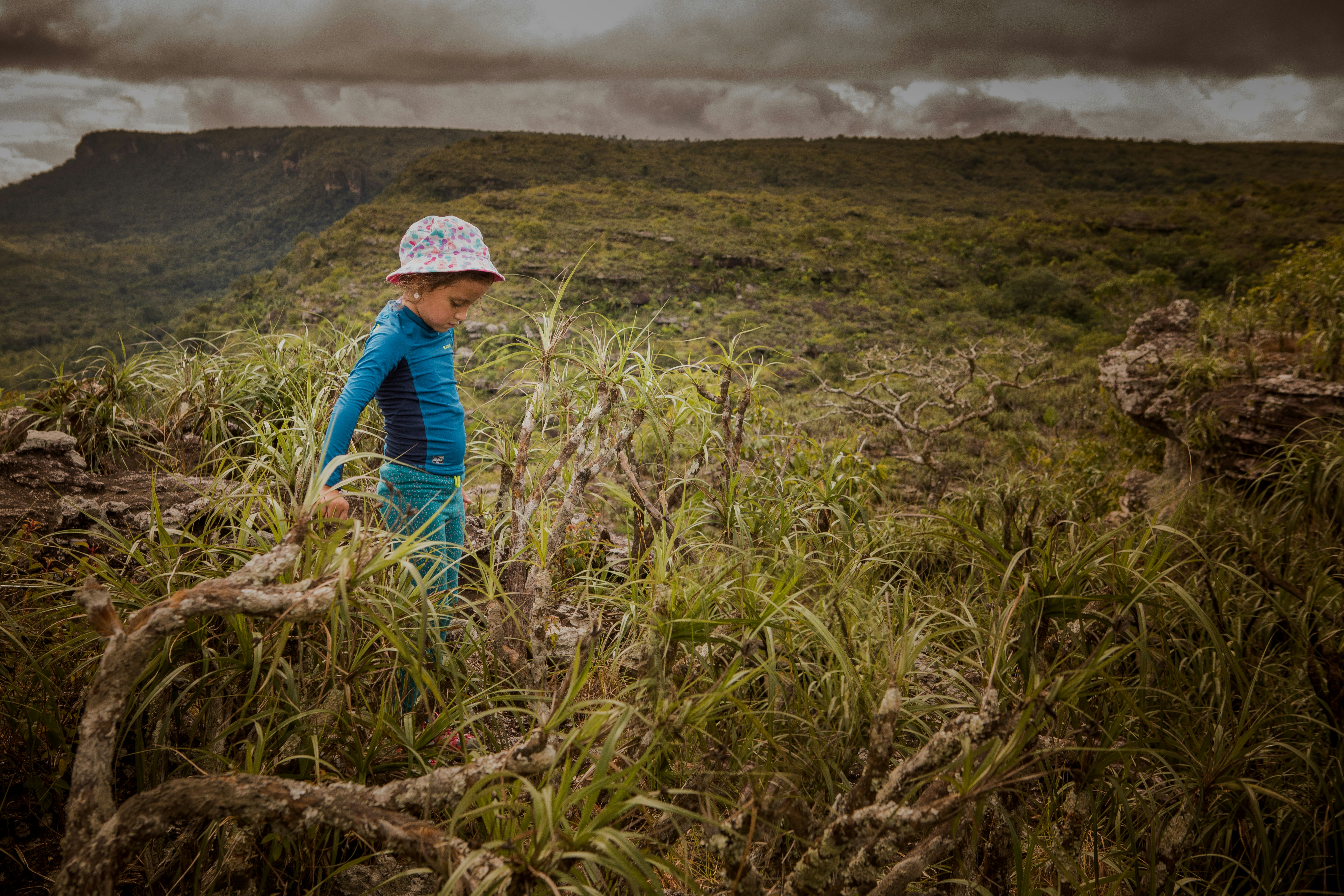 A little girl in a pink and white bucket hat and a blue rash guard stares down at the ground in front of her as she stands amidst the thick vegetation near Cano Cristales in Colombia